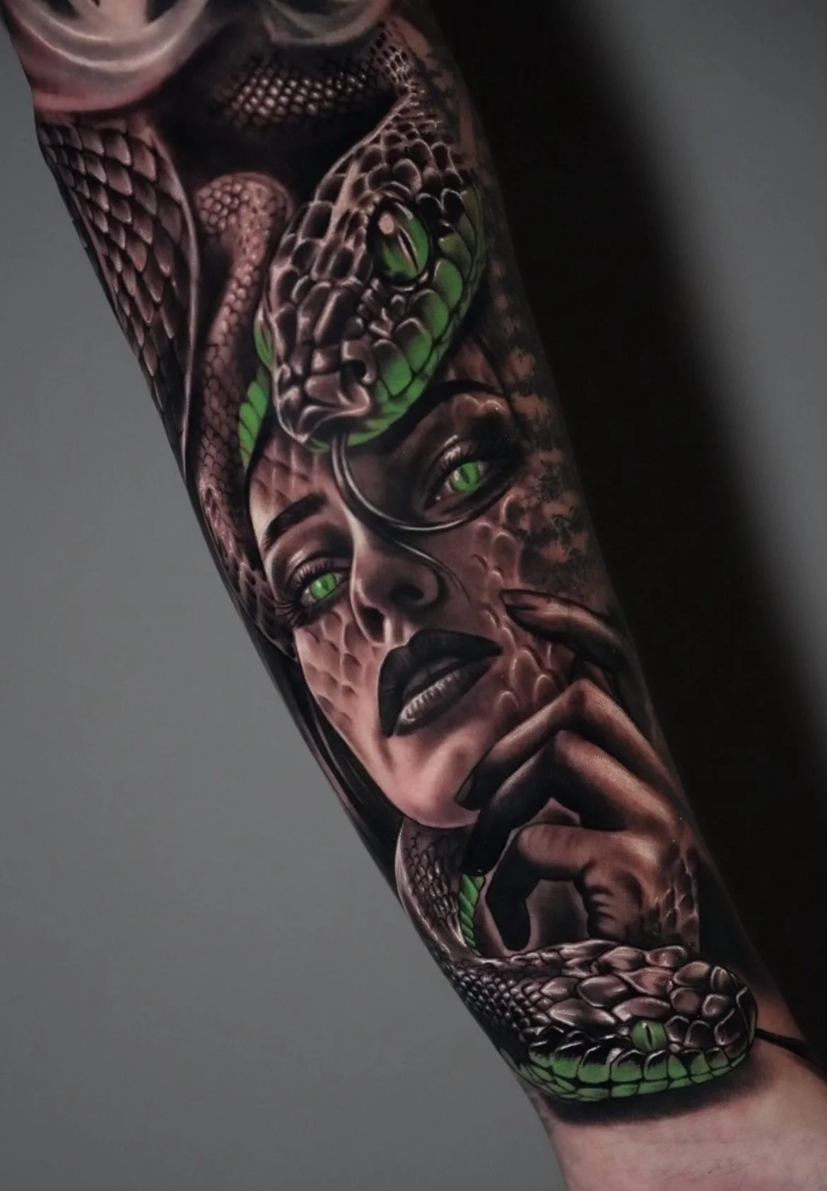 Tattoo of a woman's face with green eyes, dark lipstick, and finger on lips, with a snake coiled around her head and neck, featuring green eyes and detailed scales.
