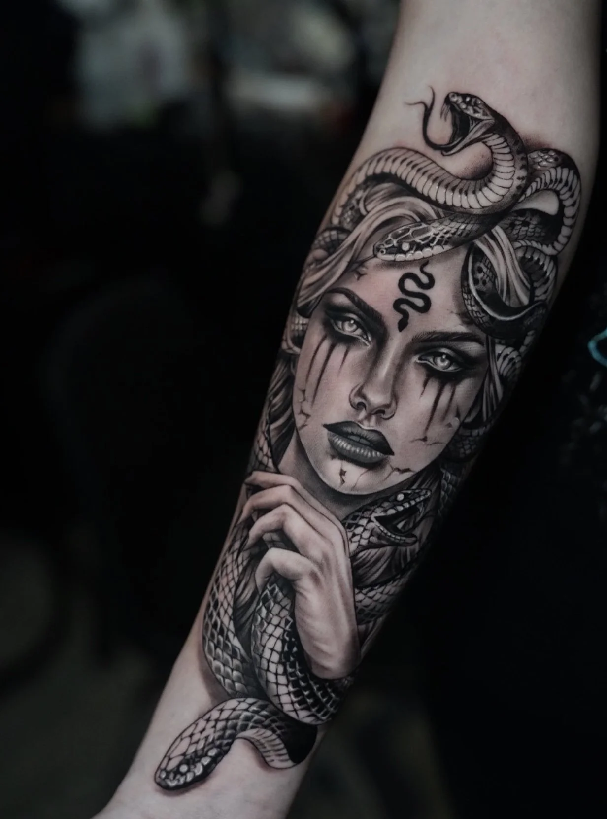 Black and gray tattoo of a woman with tears and snakes on her head, holding her chin with her hand.