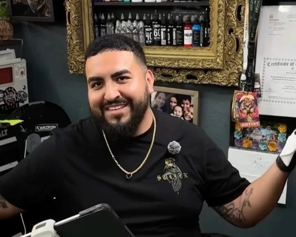 A man with a beard and short dark hair smiling at the camera, wearing a black shirt and gold necklace, in an art or tattoo studio. Behind him are framed photos, a gold ornate mirror, and shelves with ink bottles and art supplies.