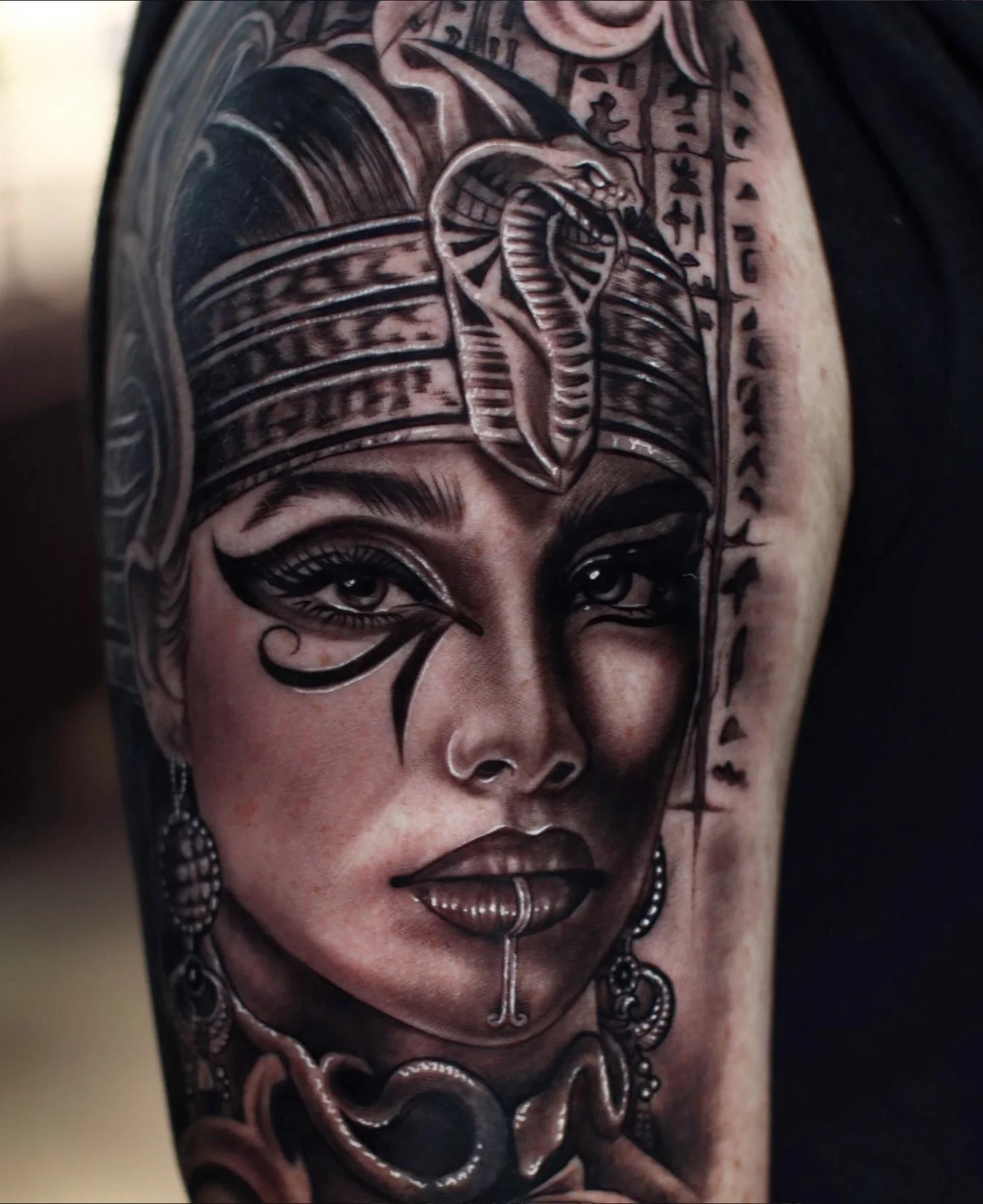 A detailed black and gray tattoo of a woman's face with traditional decorative elements, including a snake on the forehead and jewelry, covering the shoulder.