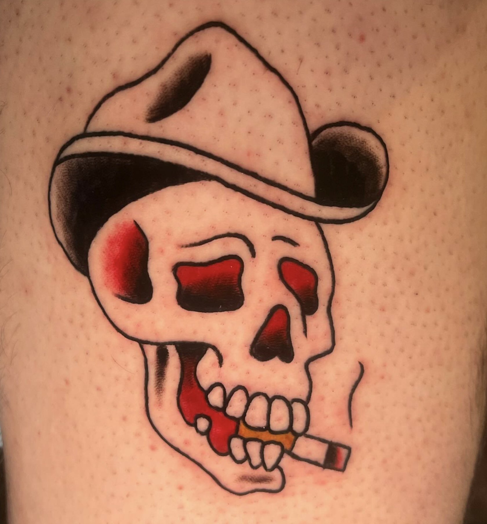 A tattoo of a cartoon-style skull with red and black accents, outlined in black, on light skin.