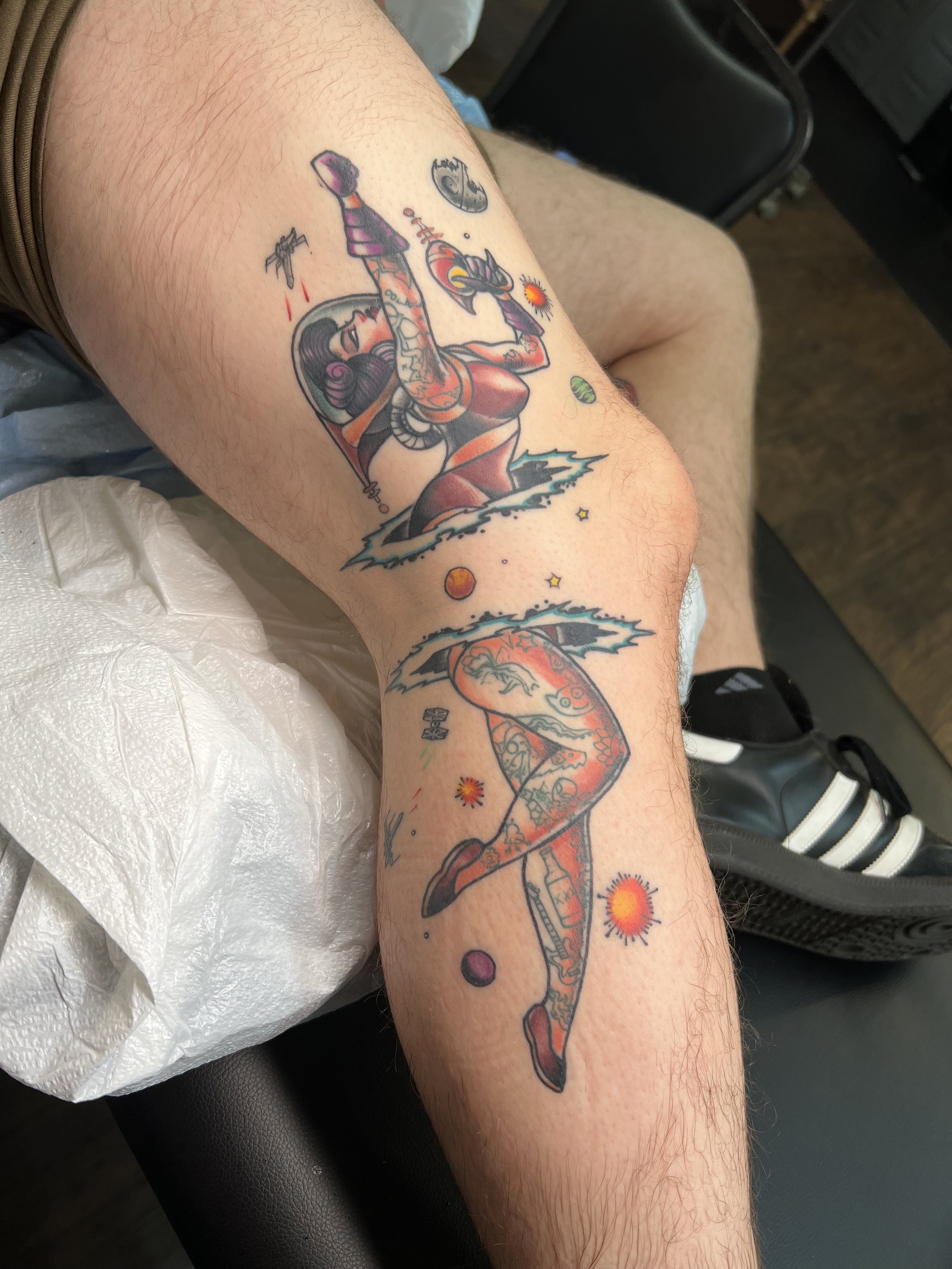 Tattoo of a pin-up girl with long dark hair and red dress, holding a space gun, surrounded by planets, stars, and cosmic imagery on a person's leg.