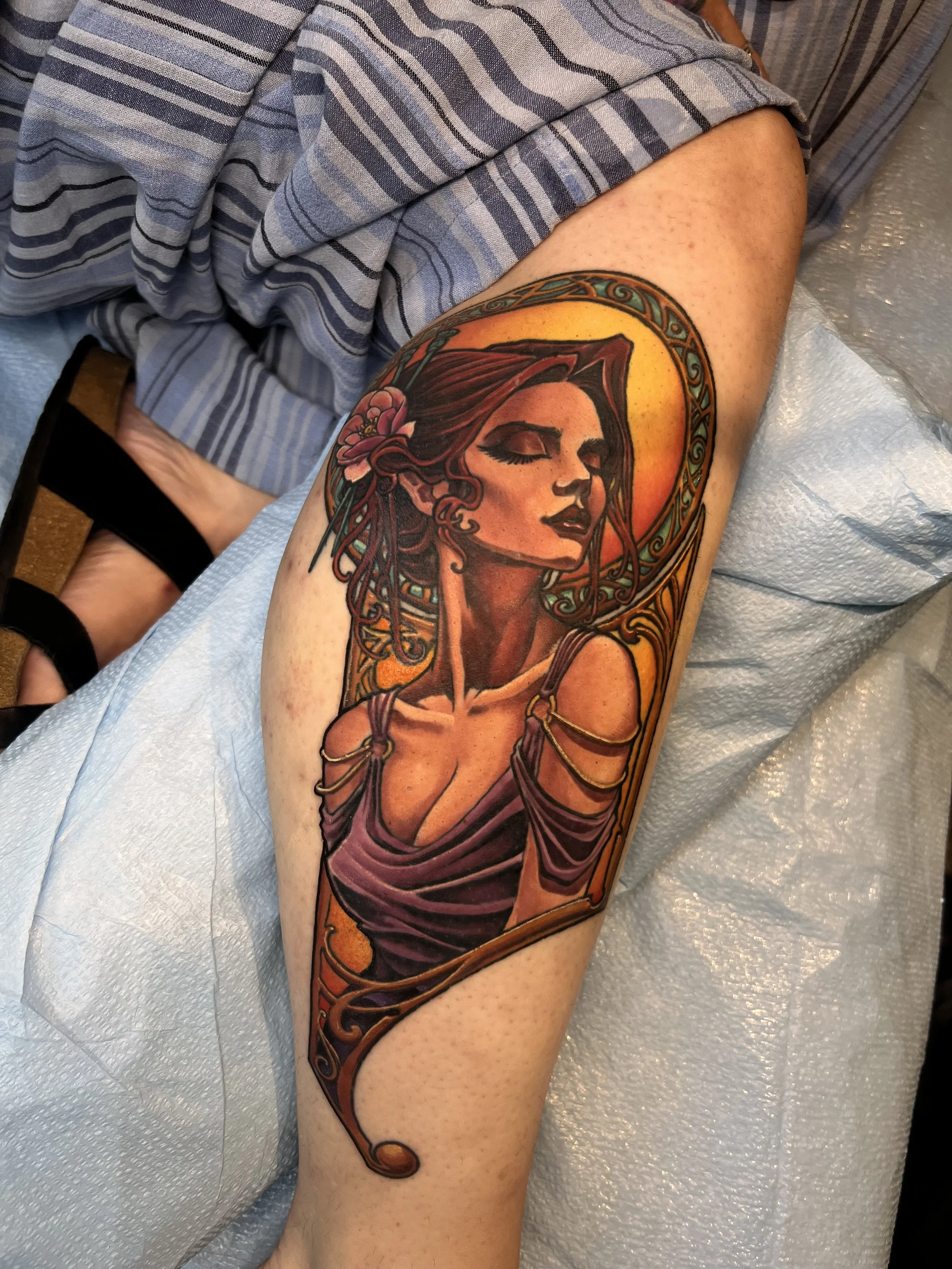Tattoo of a woman with closed eyes, long hair, surrounded by a yellow-orange halo, and floral elements on her head, on a person's upper arm.