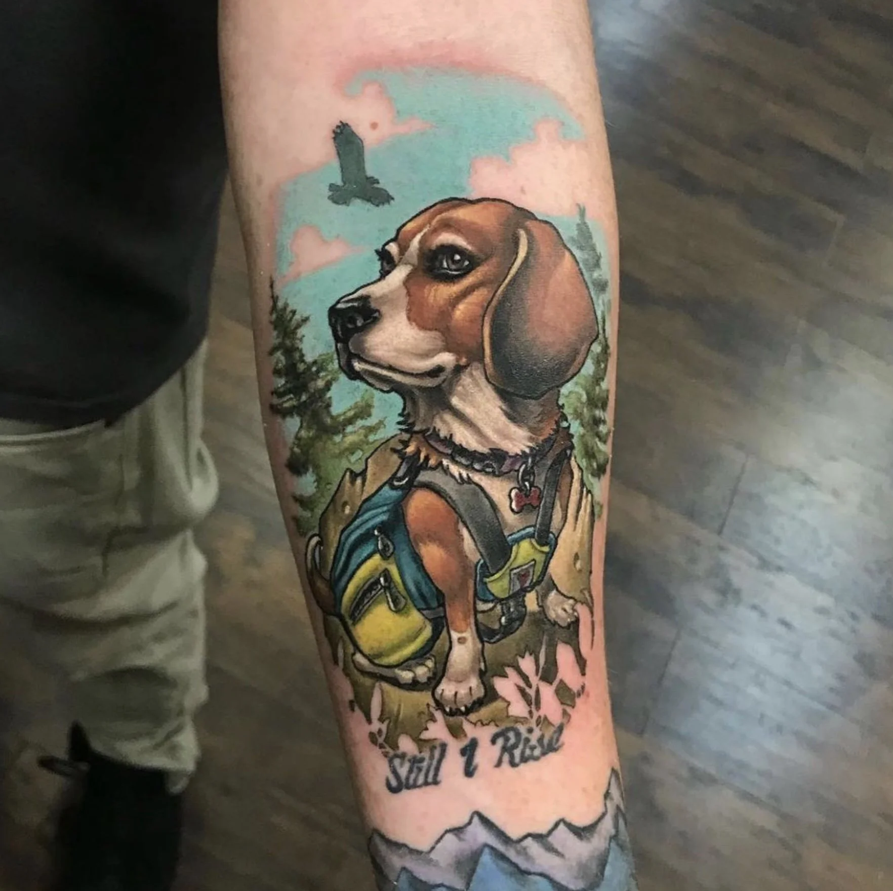 Colorful tattoo of a dog with a backpack and colorful background including a flying bird and scenic trees, with text at the bottom.