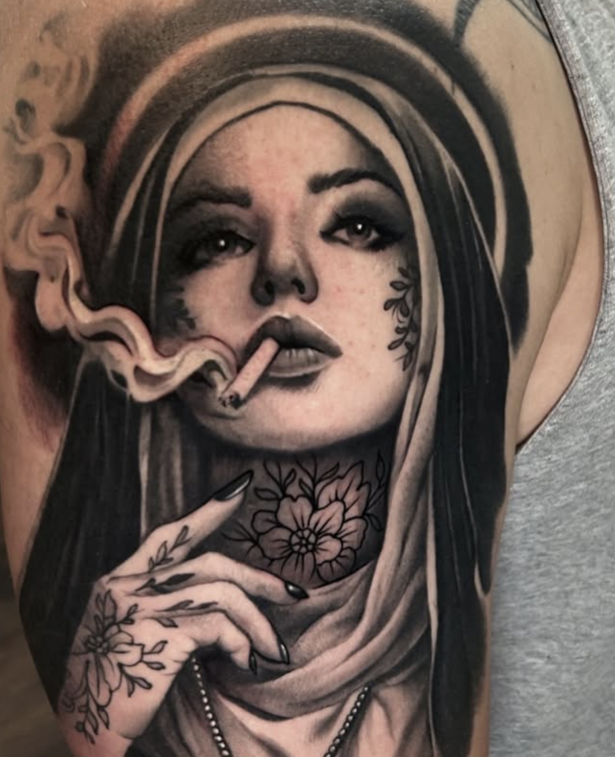 Black and gray tattoo of a woman with dark makeup, holding a cigarette, with flowers on her neck and hand, wearing a headscarf.
