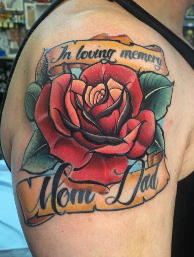 Tattoo of a red and pink rose with green leaves, with on a banner above it that says 'In loving memory' and below that 'Mom' and 'Dad' in cursive.