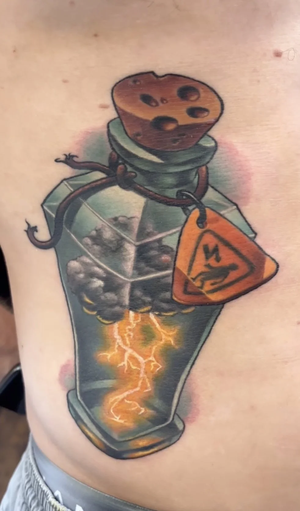 A tattoo depicting a cartoonish grenade with a cherry tomato head, sitting on a box with a radioactive hazard symbol. The grenade is connected to a lit dynamite stick with bolt-like electricity and lightning effects, and a warning sign hangs from it.