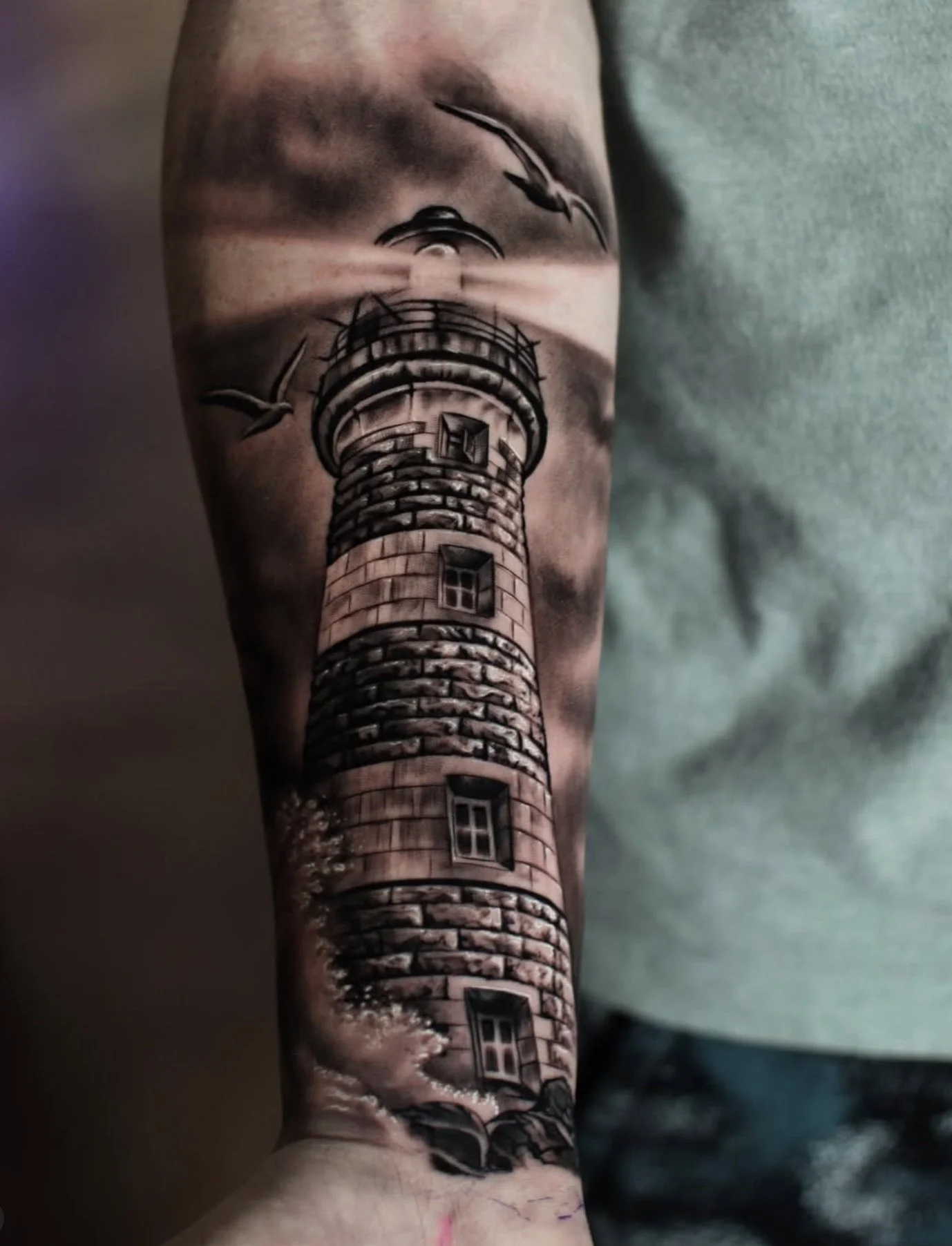 Tattoo of a lighthouse on a person's arm, with a building at the top and clouds in the background.