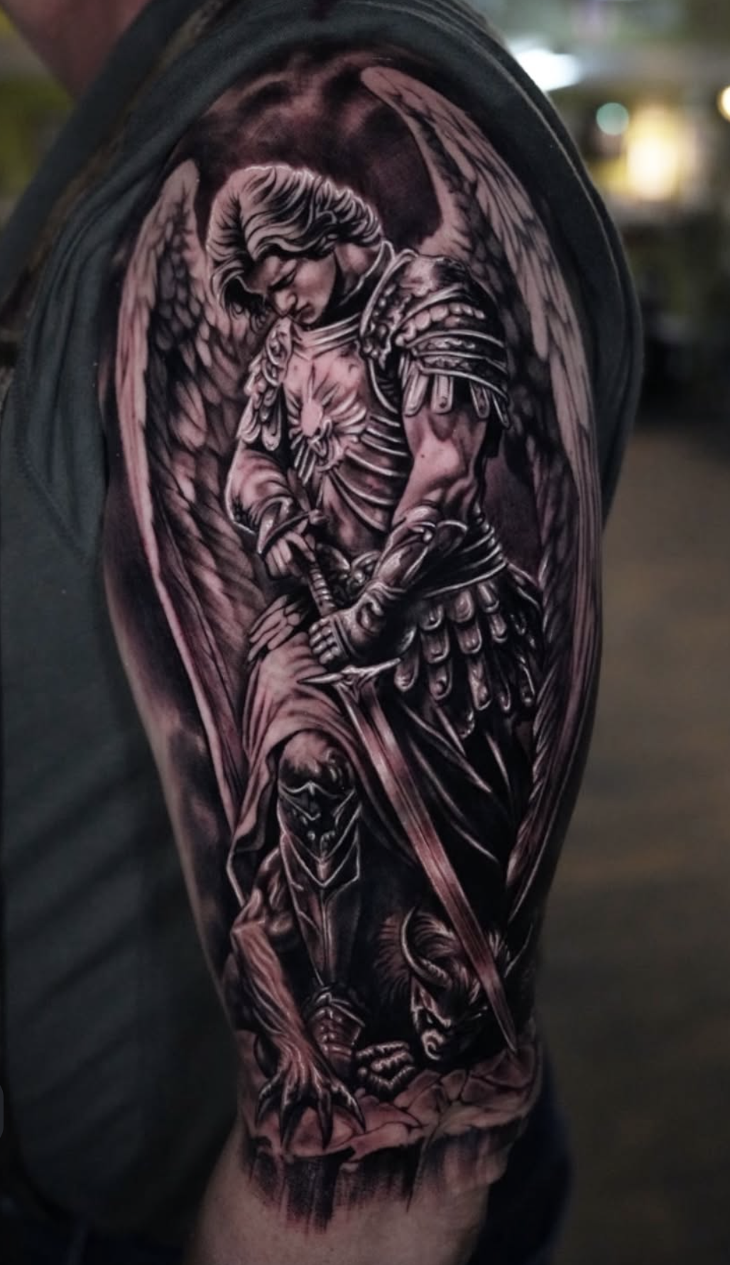 Detailed black and gray tattoo of an angel with large wings, wearing armor, holding a sword, and standing over a demonic creature.