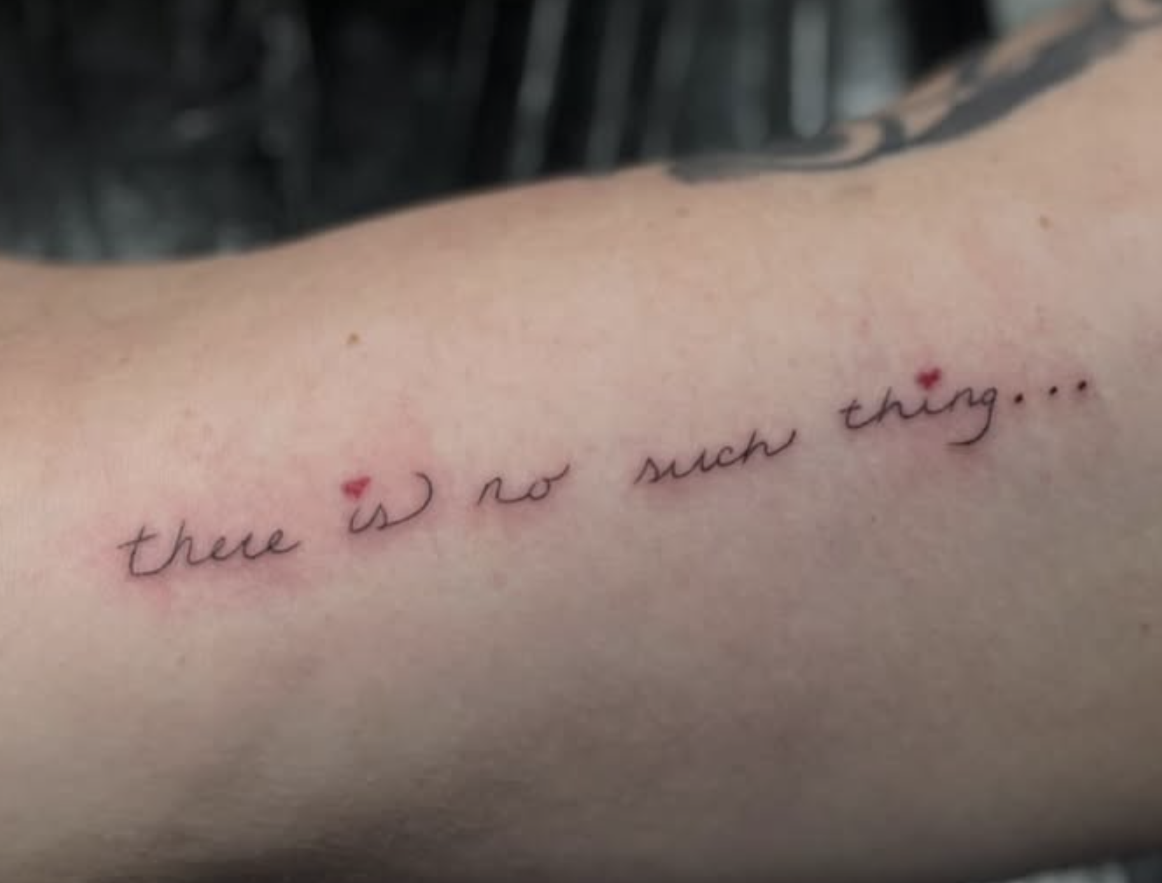 A tattoo on a person's skin with the phrase "there is no such thing..." written in cursive, featuring small red heart symbols above the words.