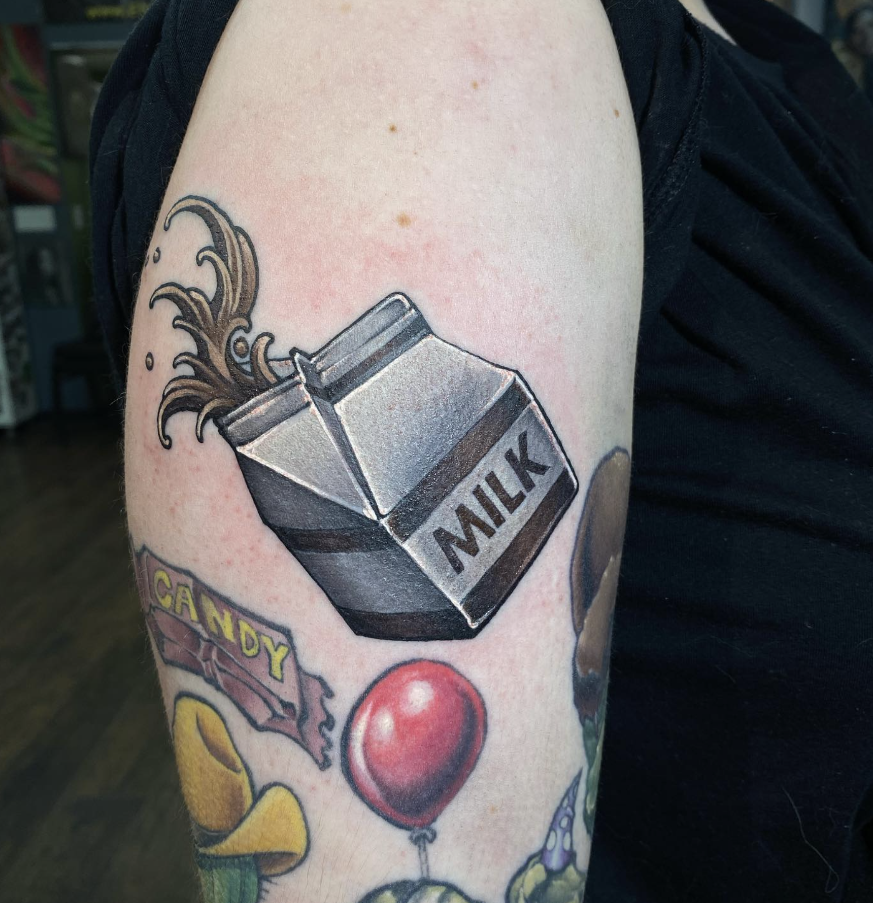 Tattoo of a milk carton with liquid pouring out, a red ball, a yellow object, and the words 'CAMPBY' visible on the arm.