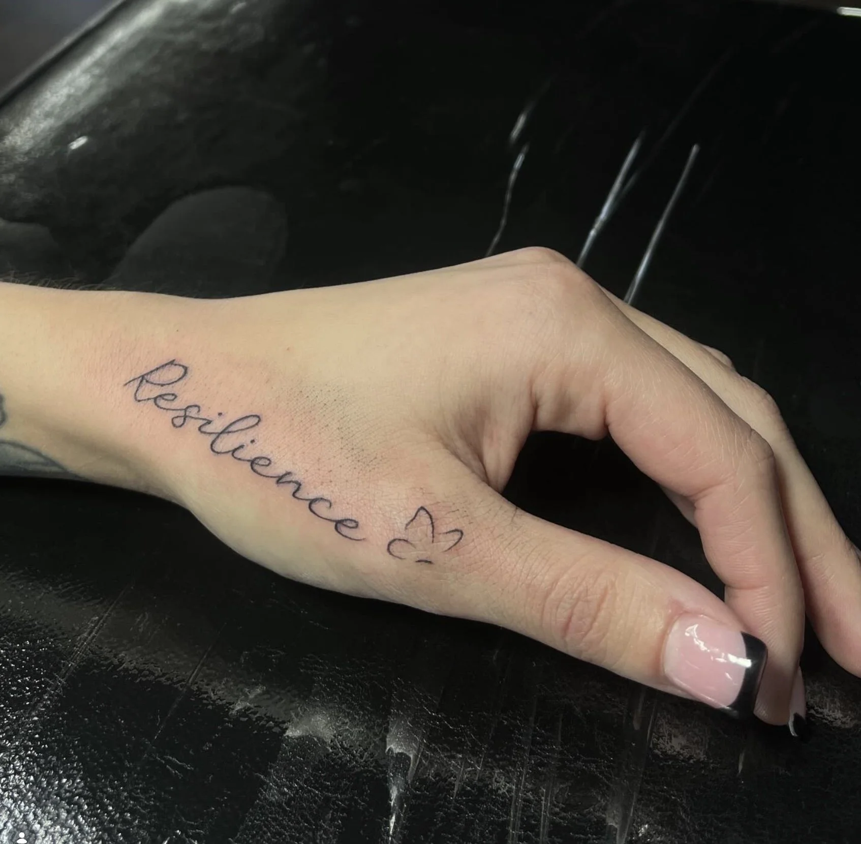 A person's hand with a tattoo reading 'Resilience' in cursive script on the inner wrist, positioned over a black surface with faint white streaks.