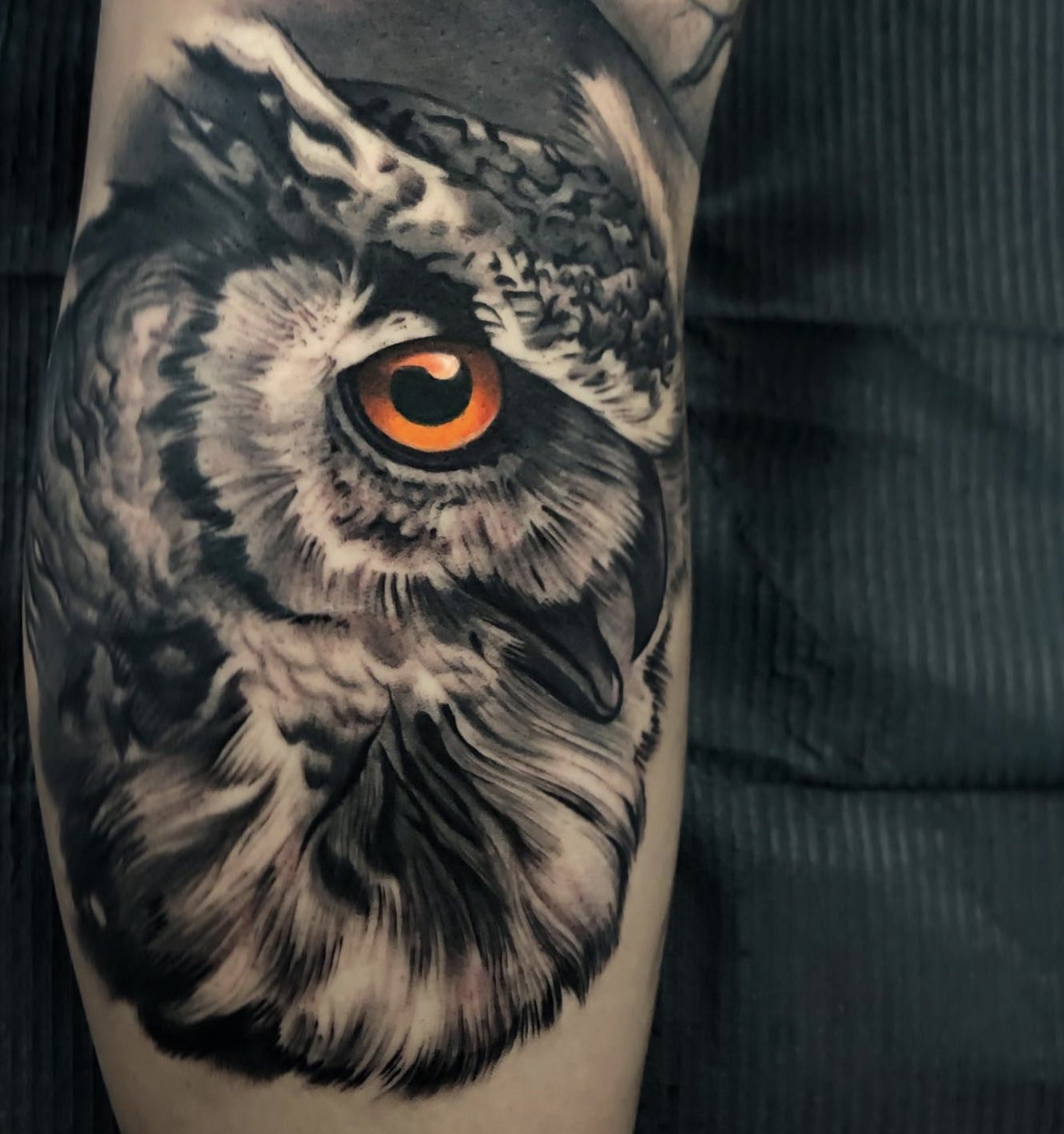 Tattoo of a realistic owl's face with intense orange eyes on a person's upper arm.