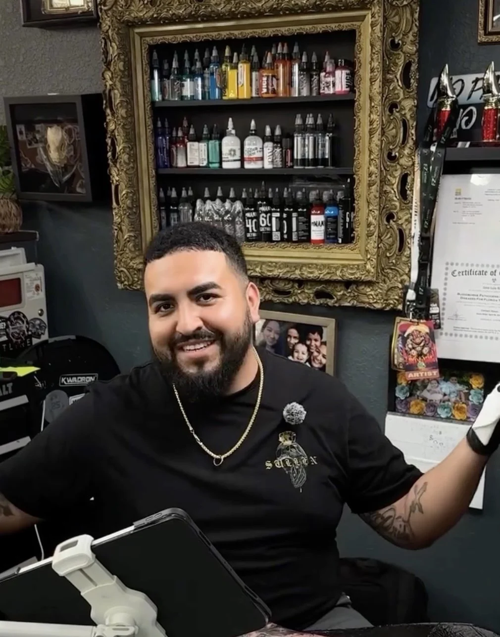 Man with a black shirt smiling with tattoo ink and pictures in the background.