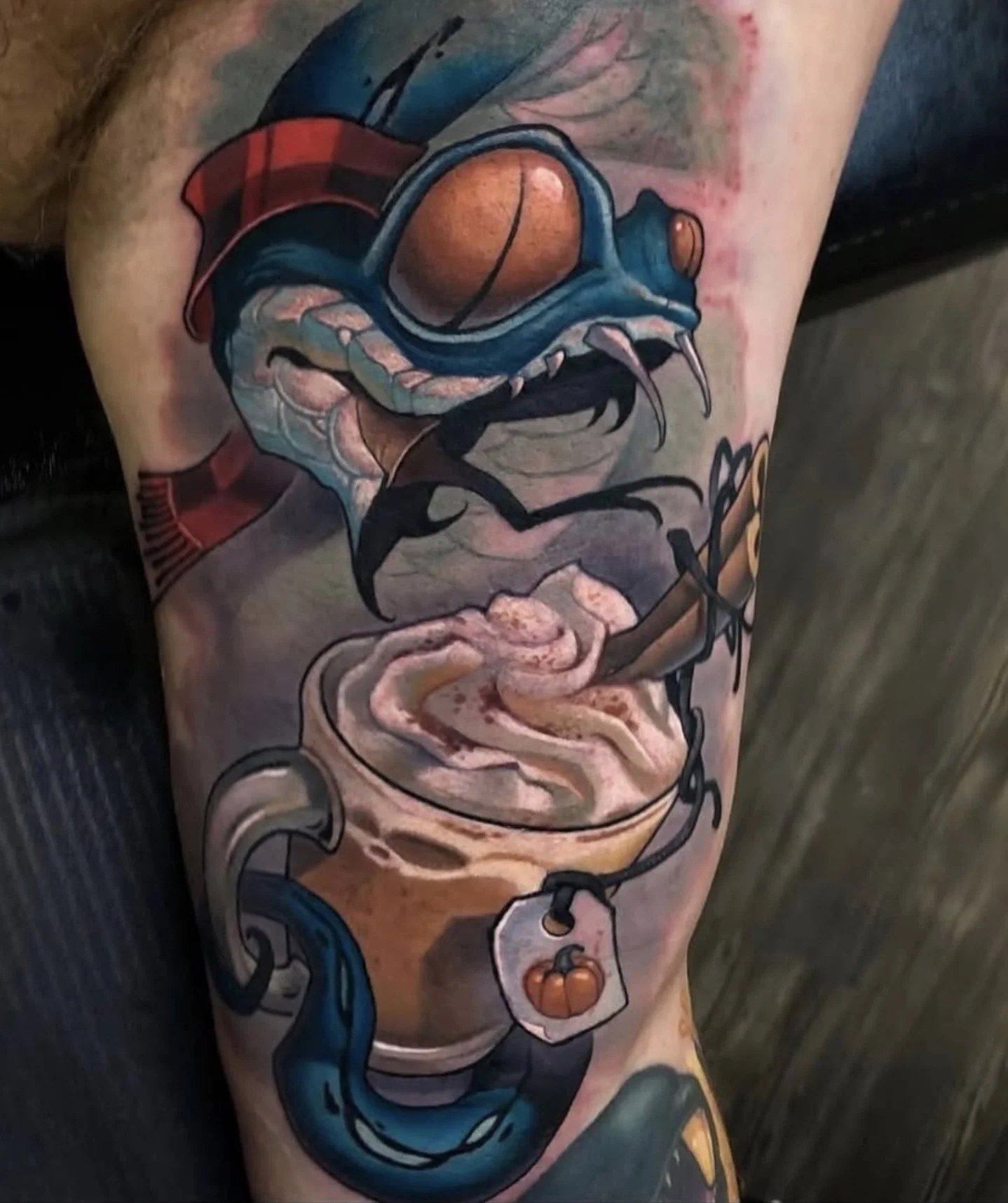 A tattoo of a stylized cartoon snake with a blue body, orange eyes, and sharp fangs, coiling above a cup of coffee with foam art, a tea bag hanging from the side, and a steam rising from the drink.