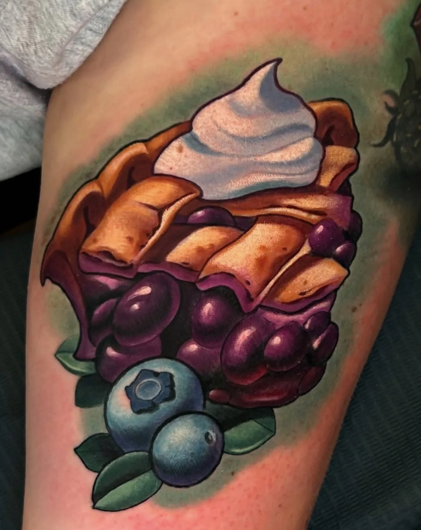 Colorful tattoo of a sliced blueberry with a vibrant, artistically rendered purple and green background on a person's upper arm.
