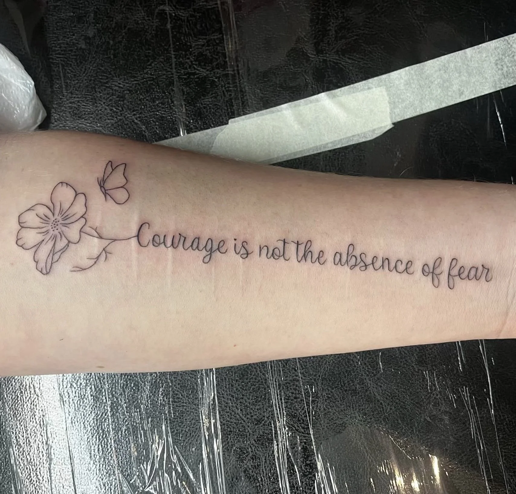 A tattoo on a person's forearm featuring a flower and two butterflies, with the quote 'Courage is not the absence of fear' written in cursive.