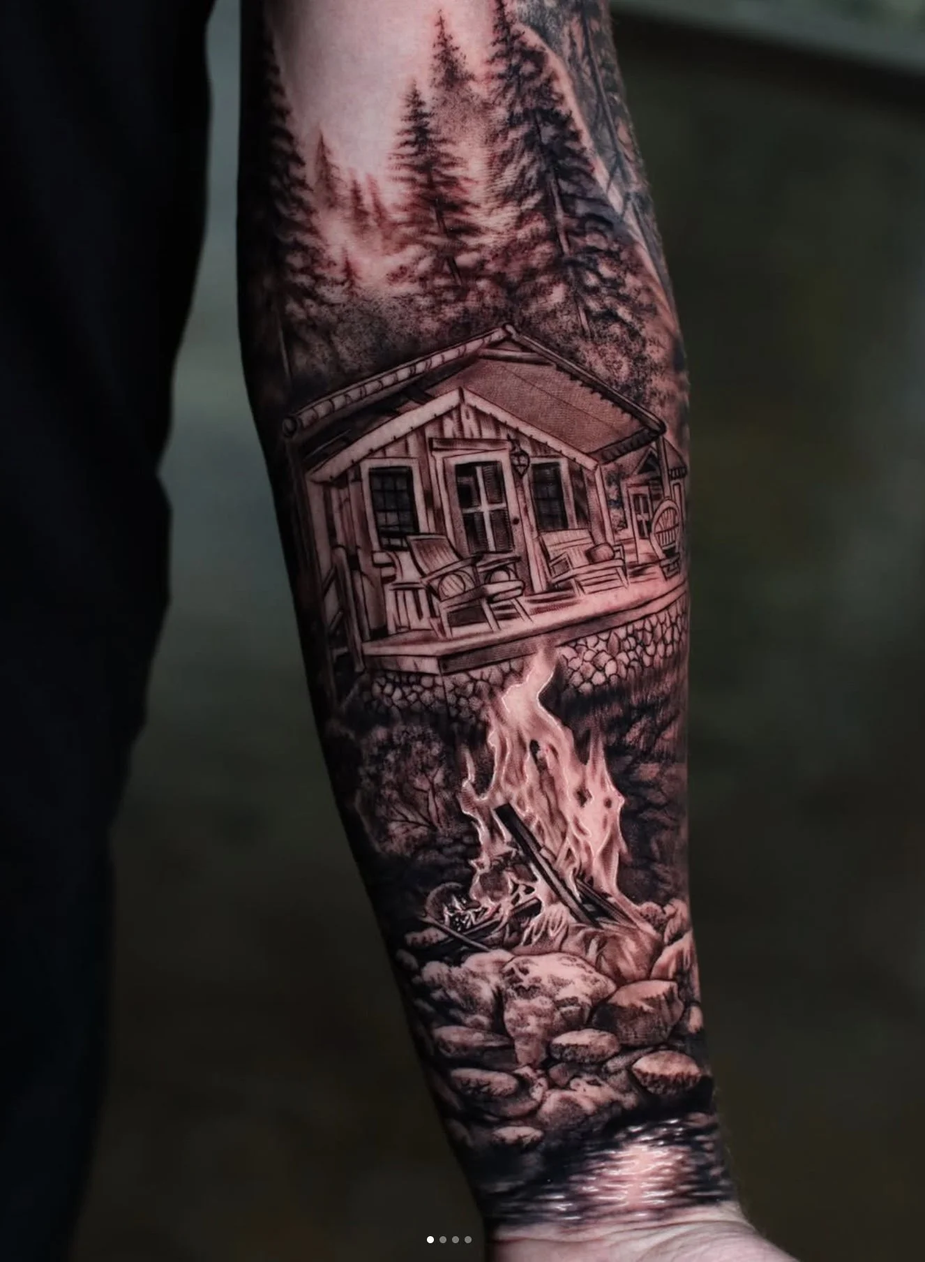 Tattoo of a cabin surrounded by tall pine trees, with mountains and clouds in the background, and a flowing stream beneath.