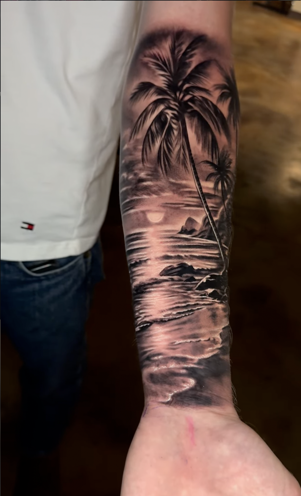 Tattoo of a beach scene with palm trees, rocks, and water at sunset on a person's arm.