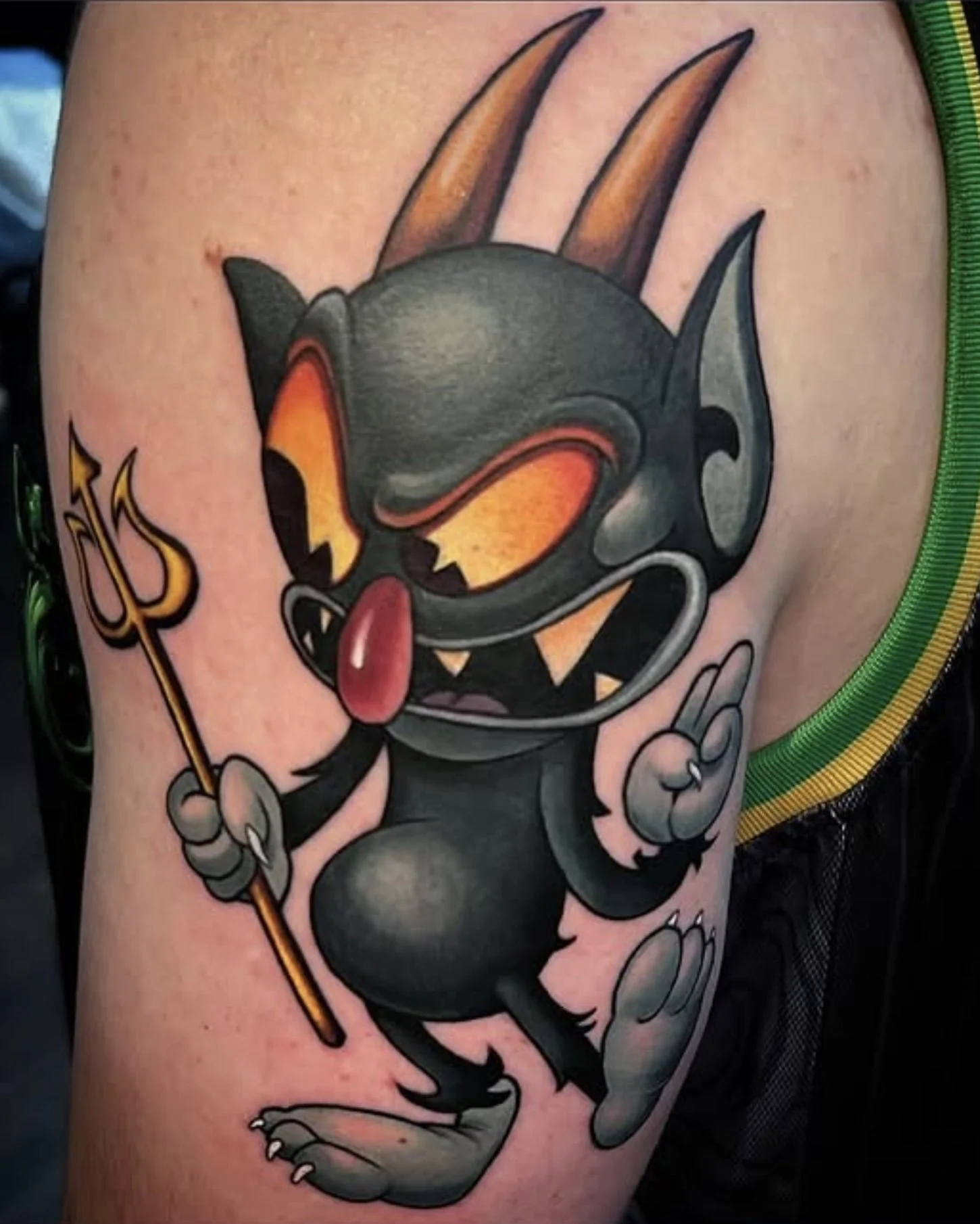 Tattoo of a cartoonish devil with orange flames on its head, holding a trident, and sticking out its tongue.