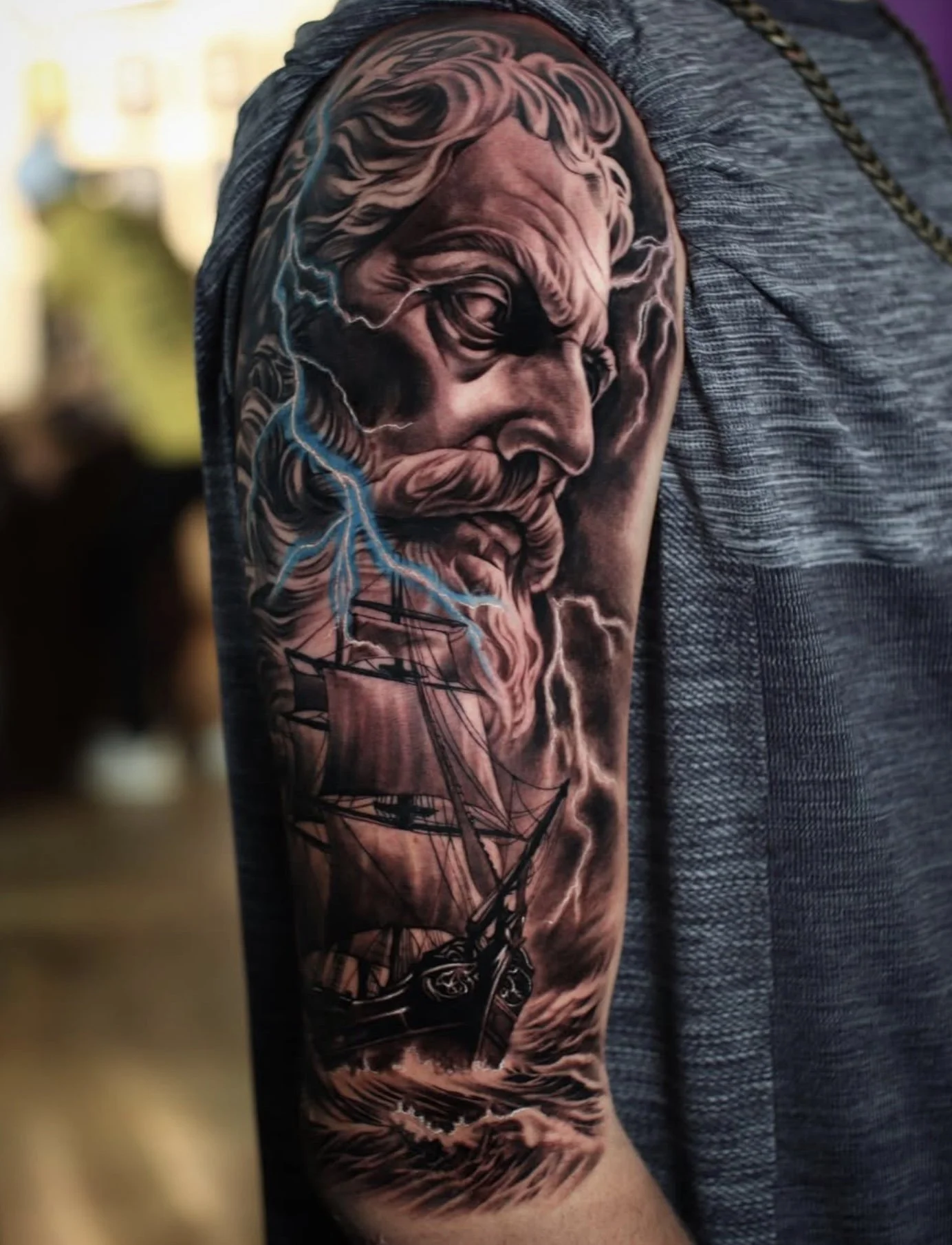 Tattoo of an elderly bearded man with flowing hair, with a ship sailing on the ocean below, and blue lightning surrounding the portrait on a person's upper arm.