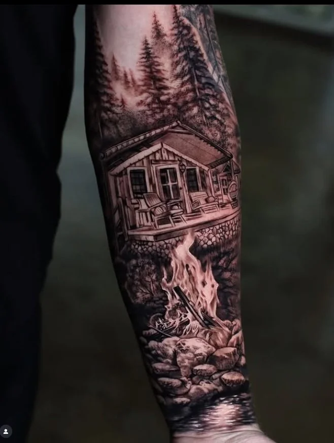 Tattoo of a cabin in the woods with trees in the background and a campfire in the foreground on a person's arm.