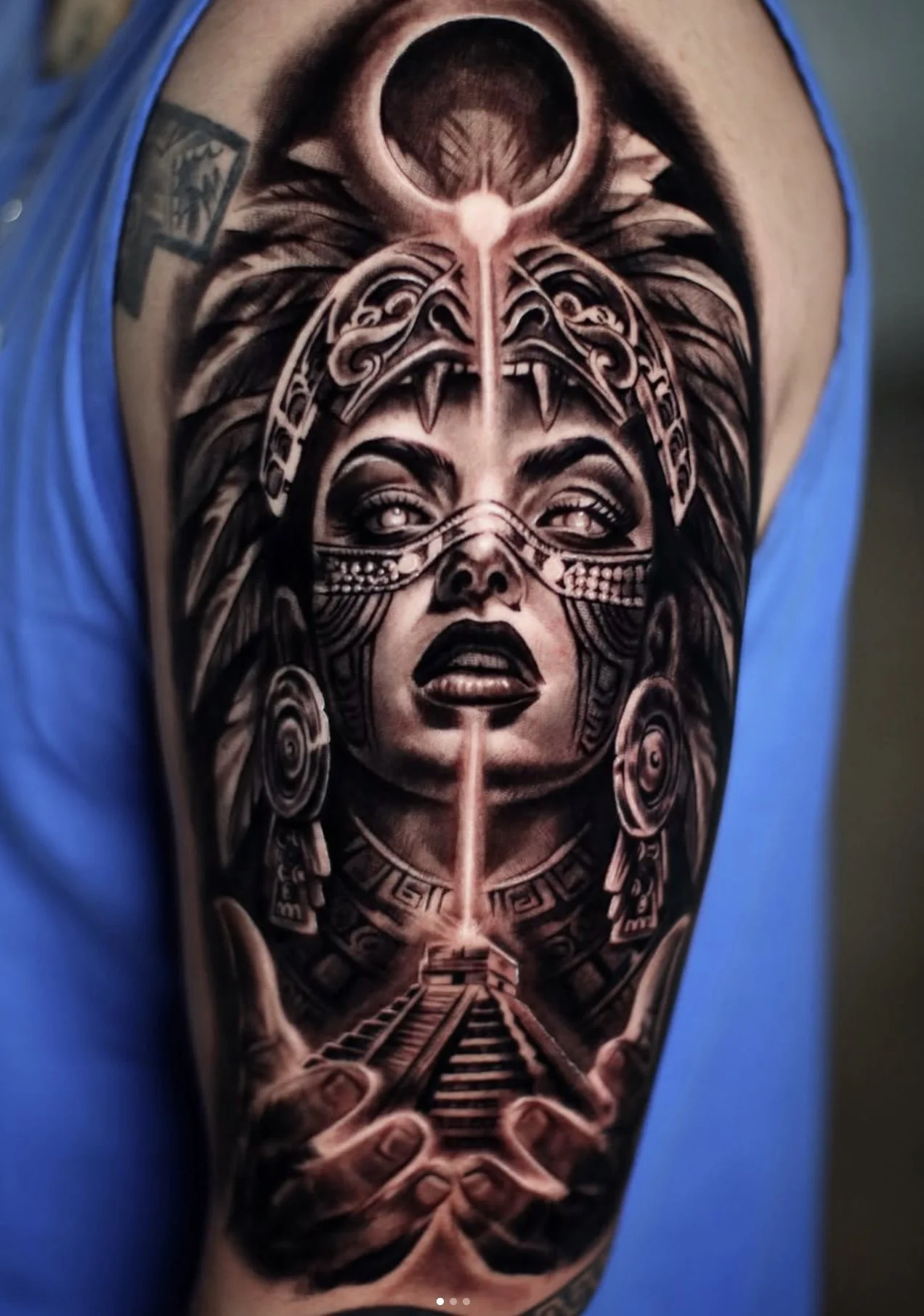 A detailed black and gray tattoo of a mystical female figure with tribal face paint, feathers, and geometric designs, featuring a pyramid with light beams from her hands.