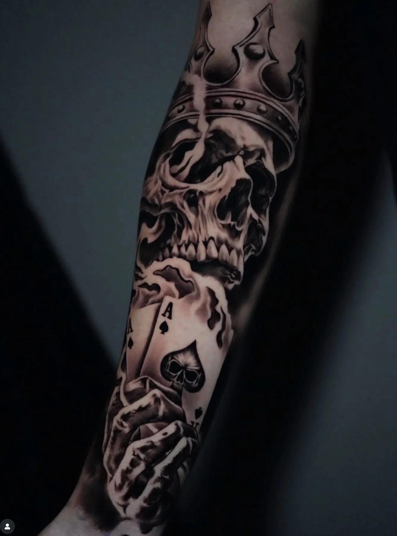 A tattoo sleeve featuring a skull with a crown, playing cards, and smoky shading, covering the arm from wrist to shoulder.
