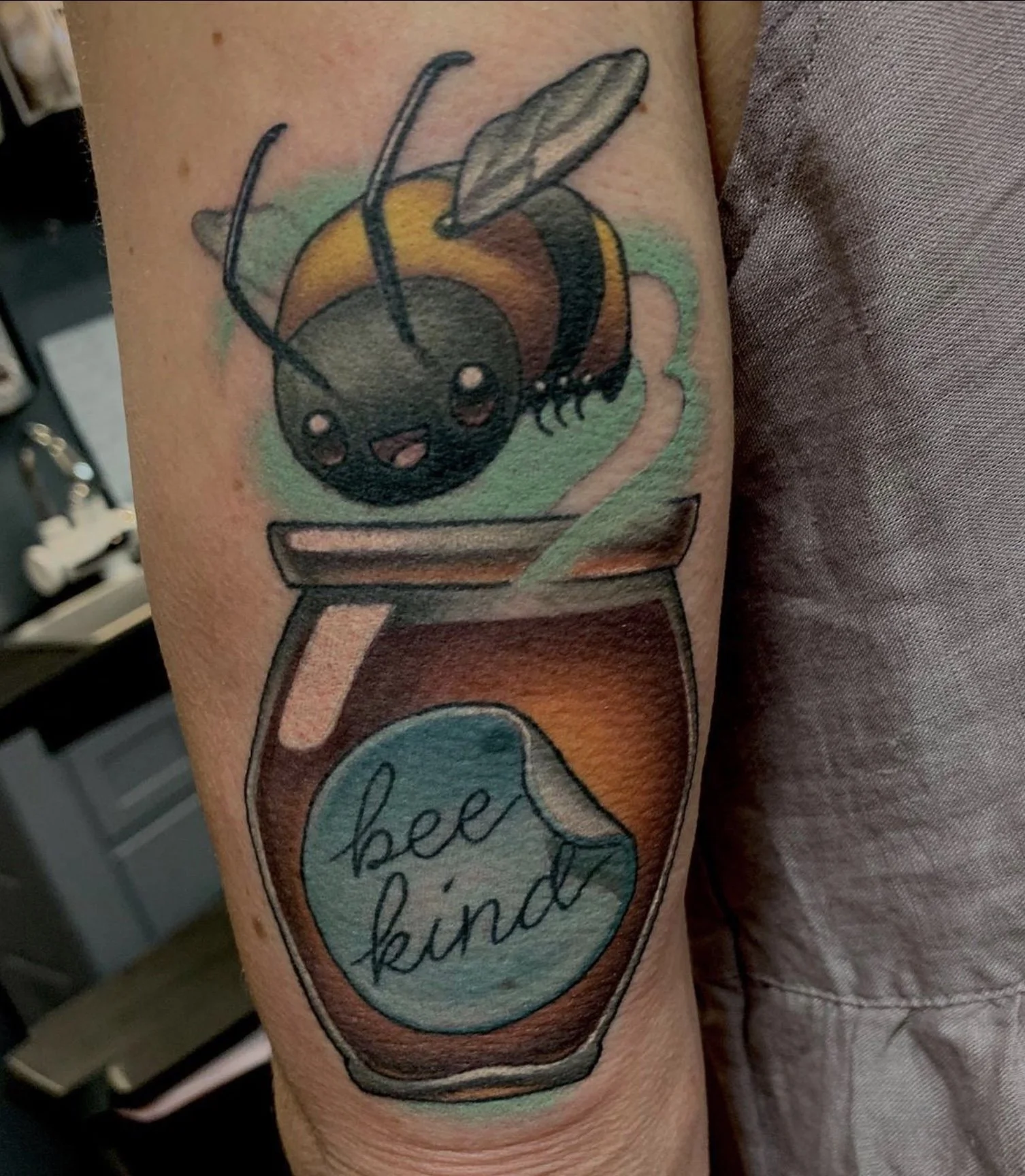 Colorful tattoo of a bee on a jar with the words "Bee Kind" in a speech bubble.
