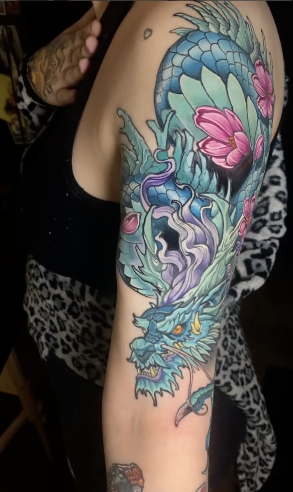 Colorful tattoo of a blue dragon with yellow eyes, sharp teeth, and an open mouth, surrounded by pink flowers and green leaves on a person's arm.