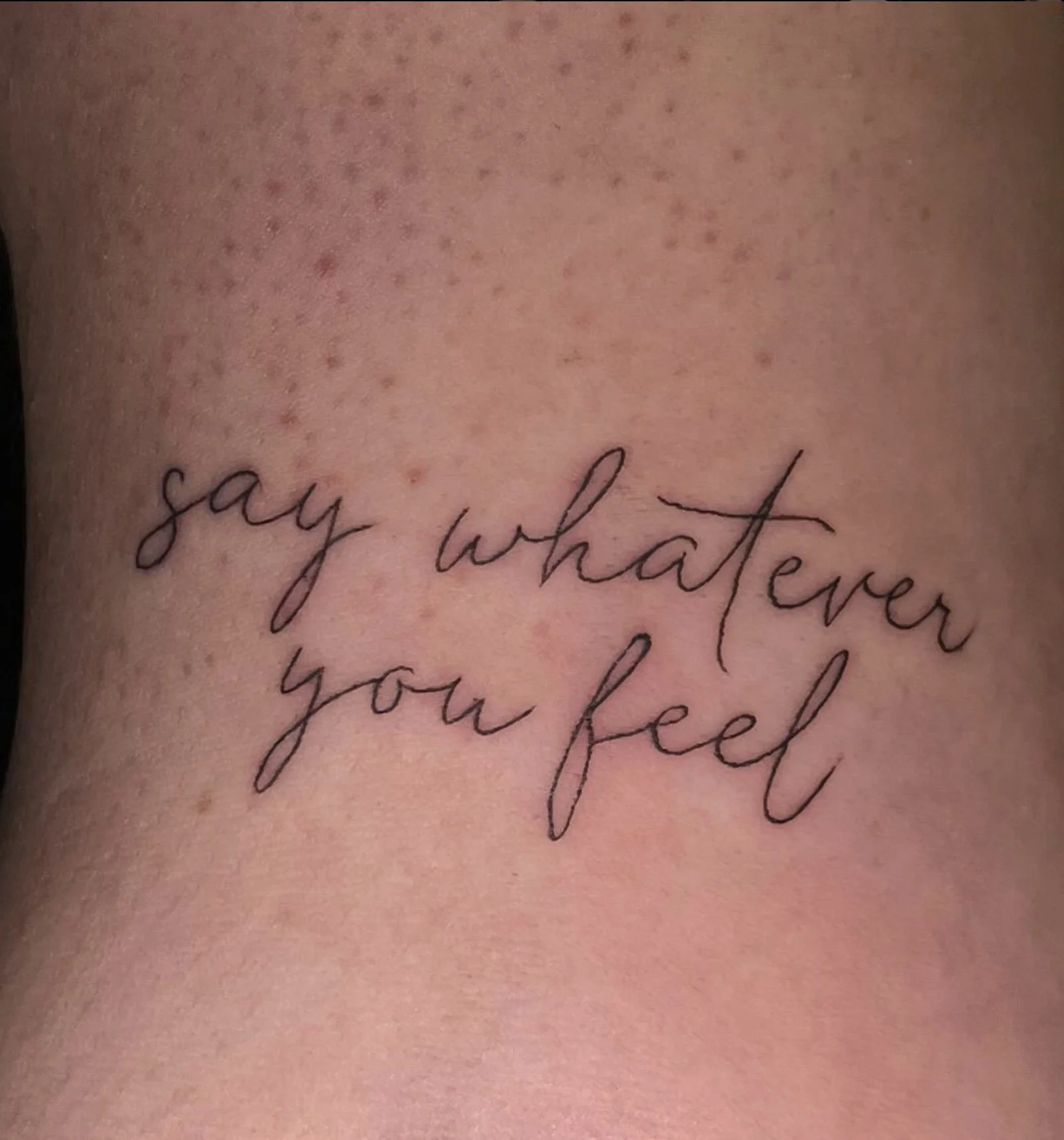 Close-up of a tattoo on skin with the words "say what ever you feel" in cursive handwriting.