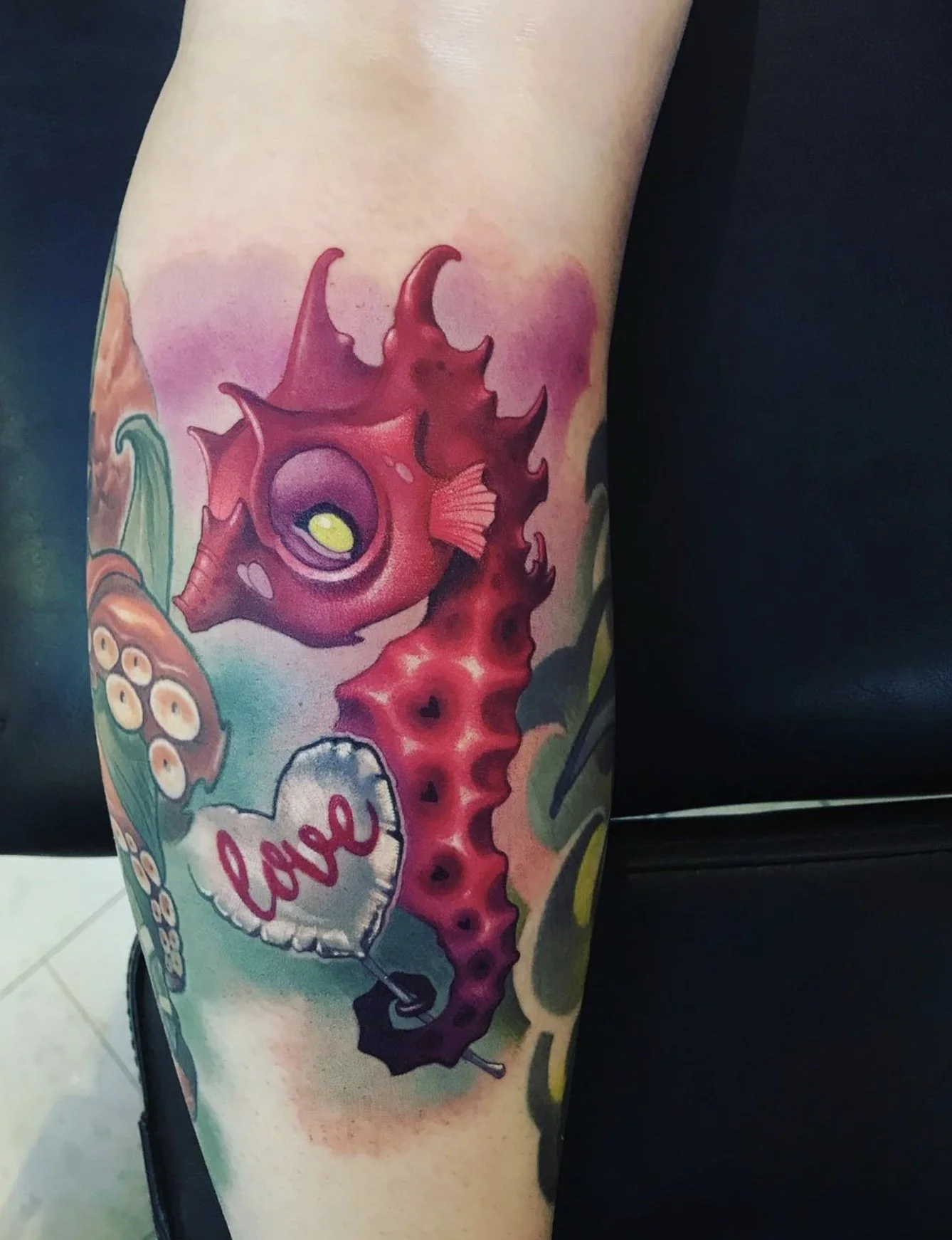 Colorful tattoo of a seahorse with pink, purple, and red hues, surrounded by ocean-themed elements including coral, bubbles, and a heart-shaped balloon with the word 'love' on it.