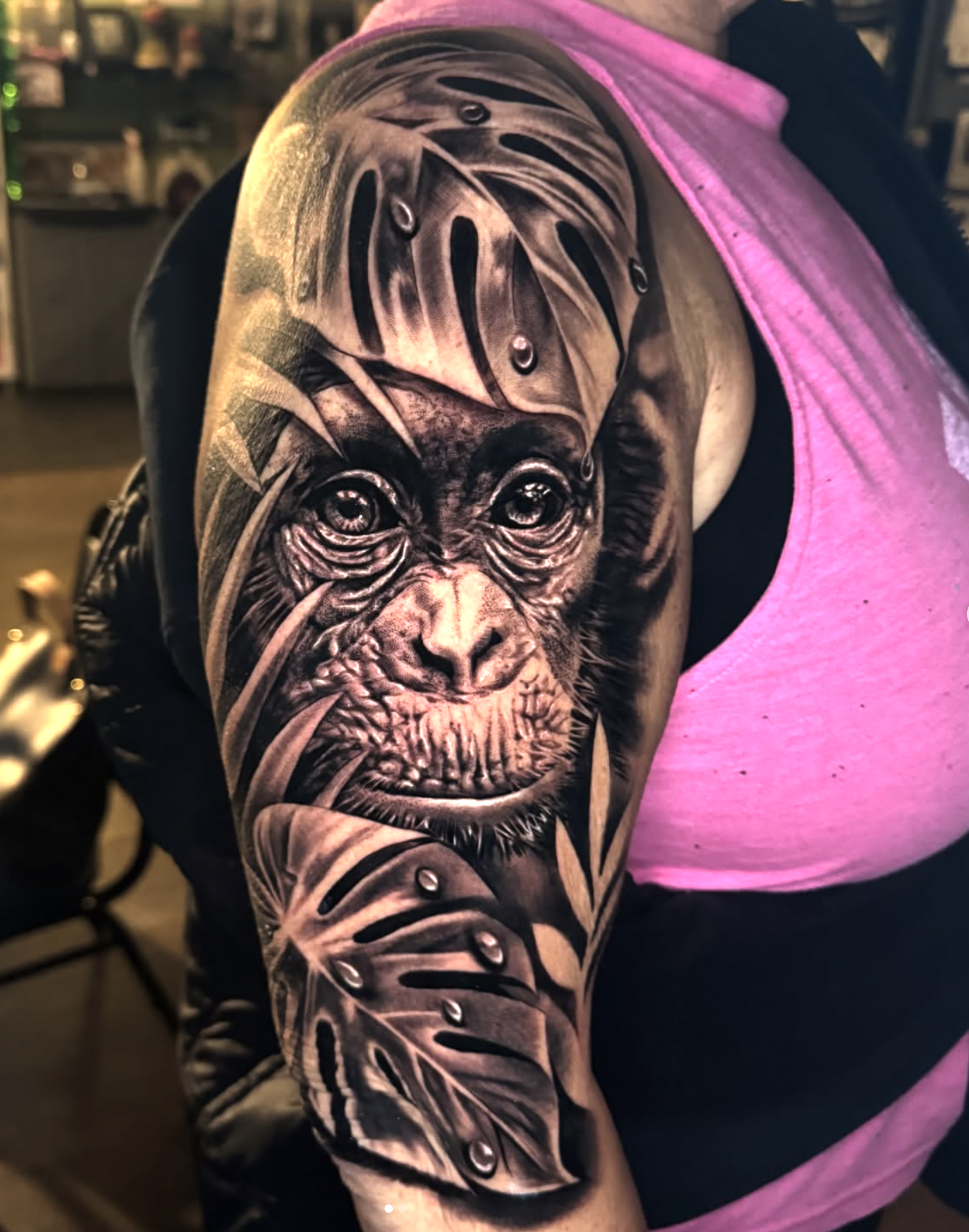 A detailed black and gray tattoo of a chimpanzee's face on a person's upper arm, with shading and realistic features, in a tattoo studio setting.