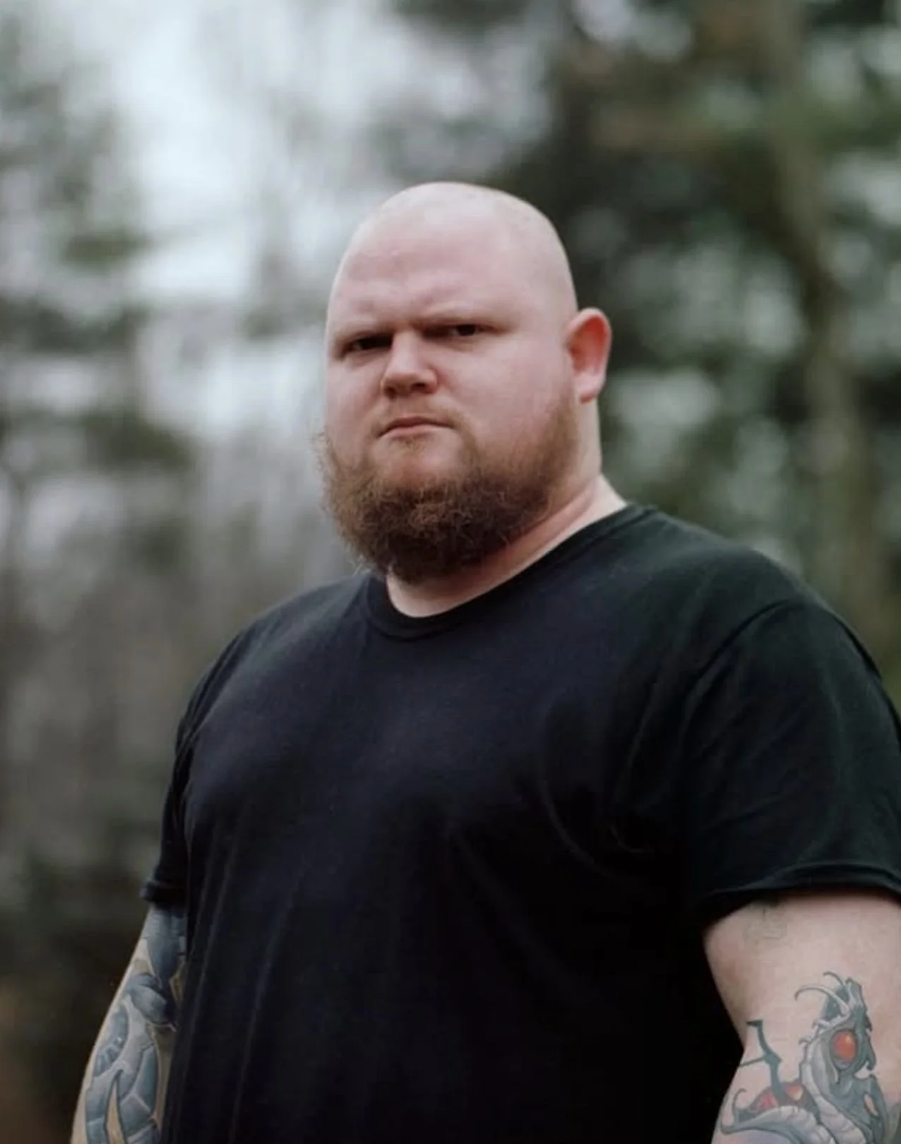 A man with a shaved head and beard wearing a black t-shirt, standing outdoors with blurred trees in the background.