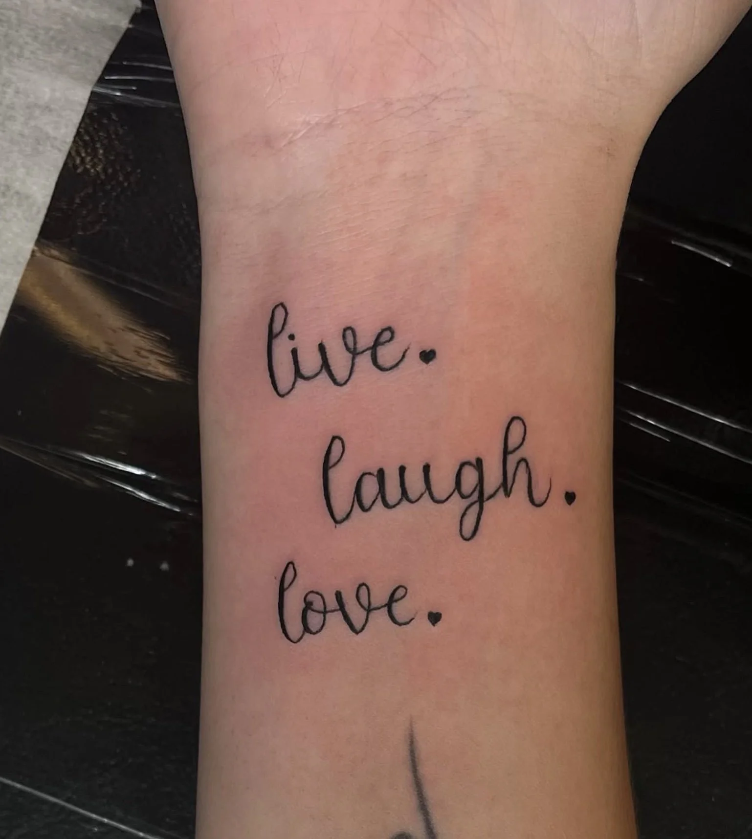 Close-up of a tattoo on a person's arm with the words "live," "laugh," and "love" written in black cursive font, each word followed by a small heart.