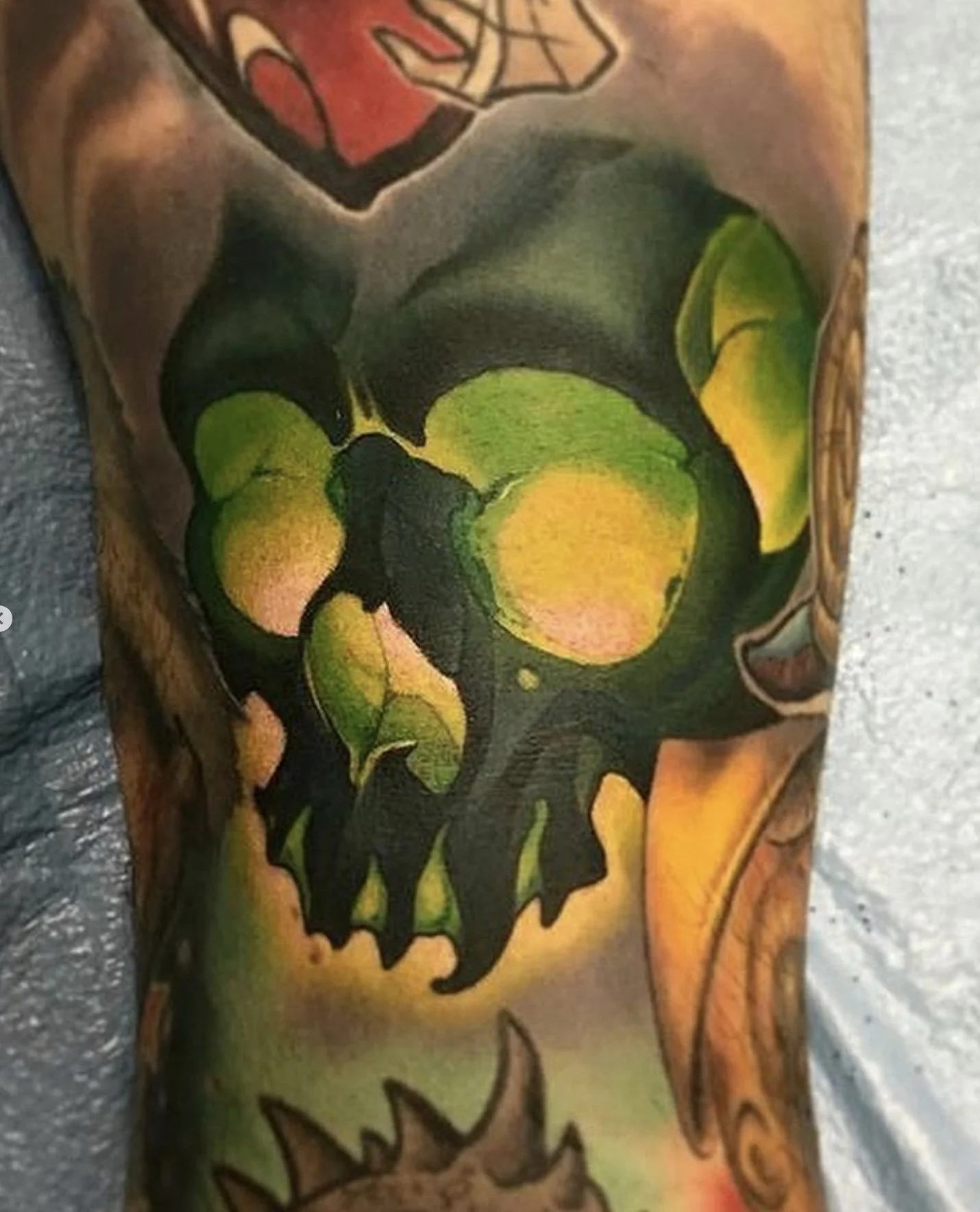 A colorful tattoo of a skull with green, yellow, and orange hues, blending into brown and black shadows, on a person's arm.