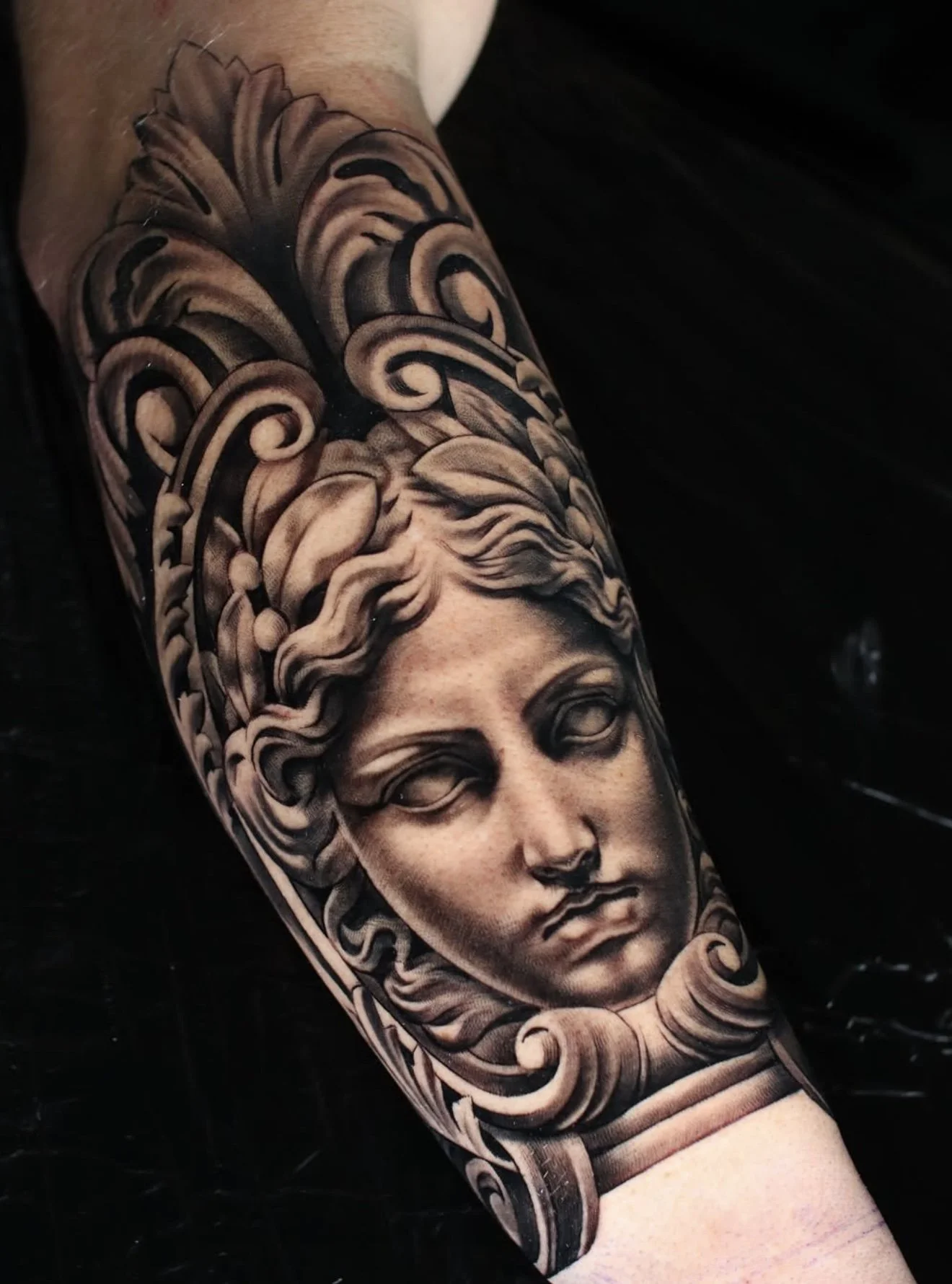 Black and gray tattoo of a classical-style woman's face with flowing hair, detailed shading, and ornamental scrollwork on an arm.