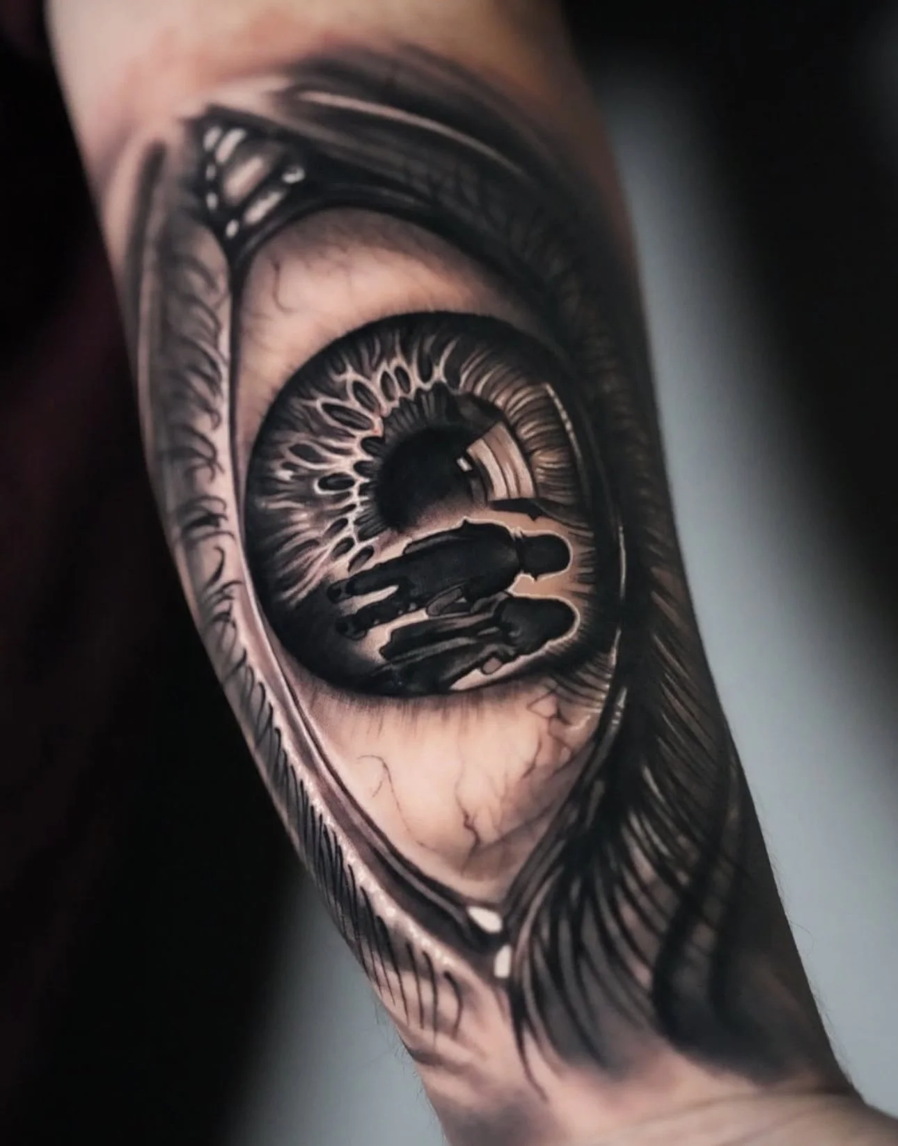 A detailed black and gray tattoo of an eye on a person's arm, with a reflection of a person walking along a street in the eye's pupil.