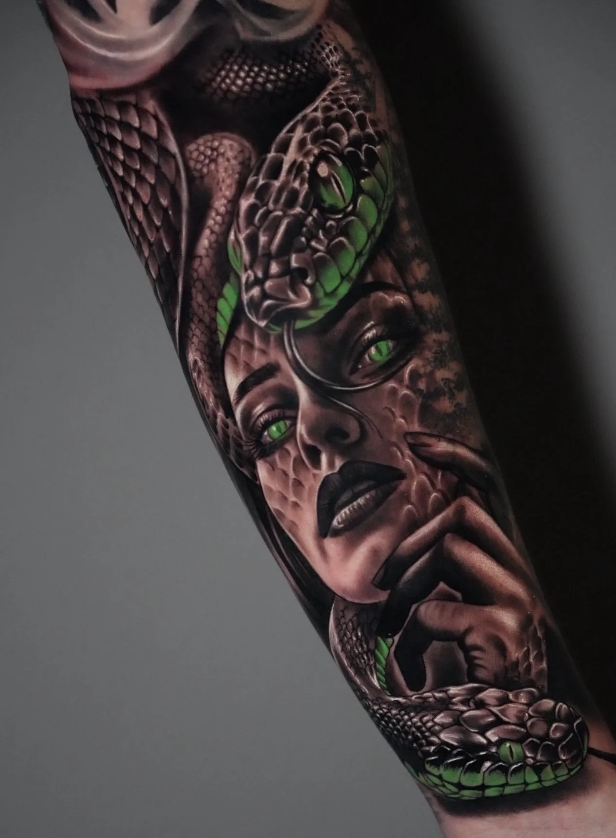 A detailed tattoo on an arm featuring a woman's face with green eyes, dark lipstick, and long black hair, intertwined with a green and gray snake.''