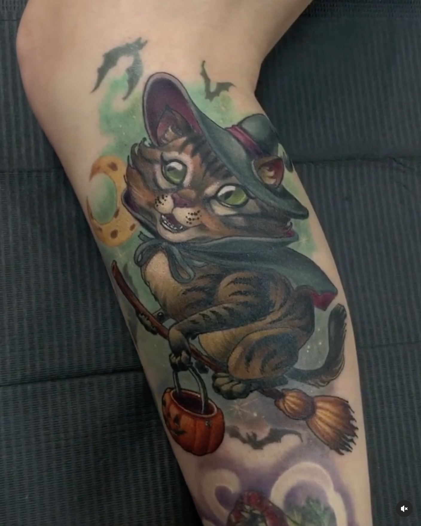 Colorful tattoo of a cat with light green eyes, holding a fishing pole with a fish on the line, surrounded by a starry night background with bats flying and a greenish moon.