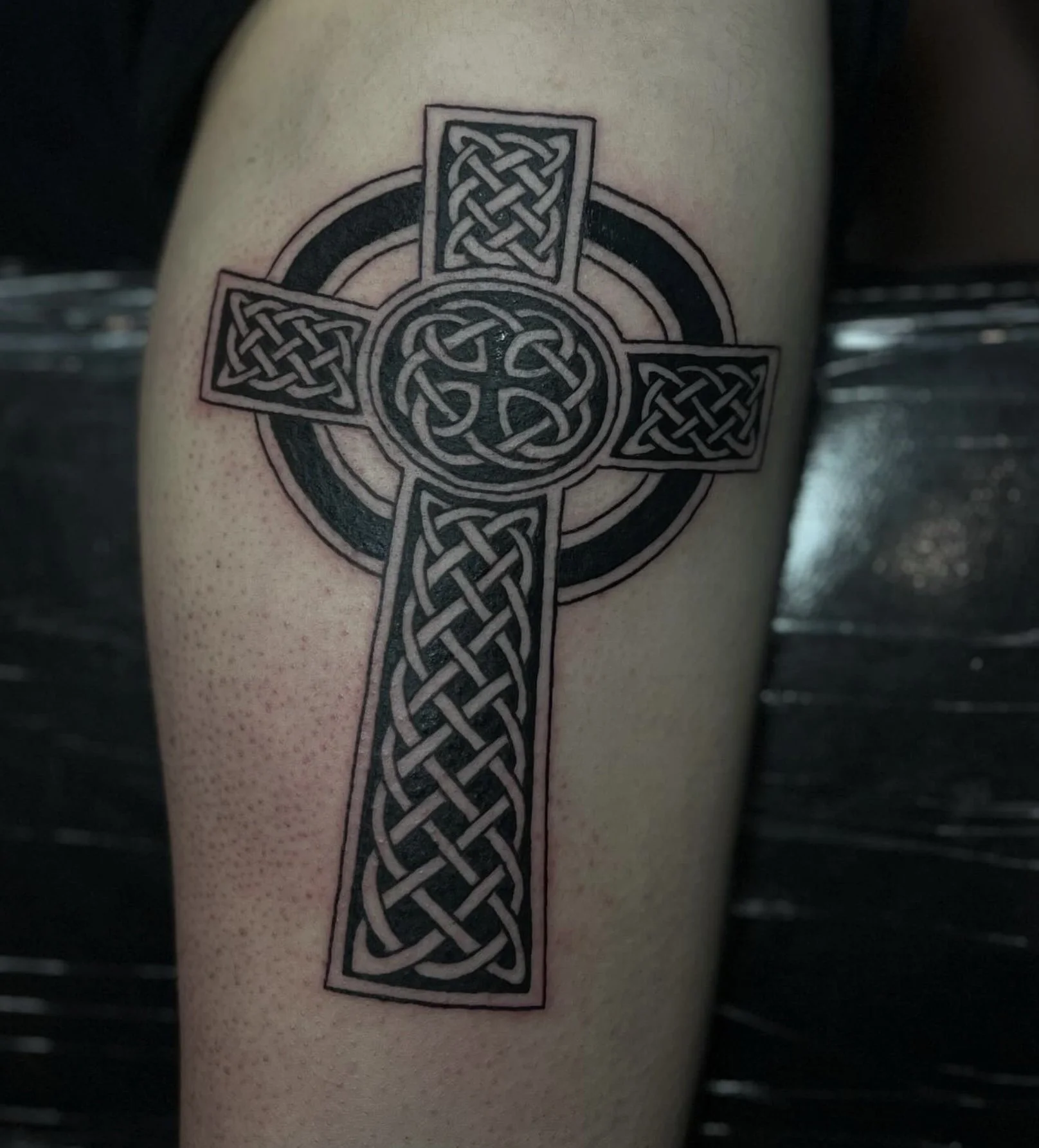 A black Celtic cross tattoo with intricate knotwork designs on a person's arm.