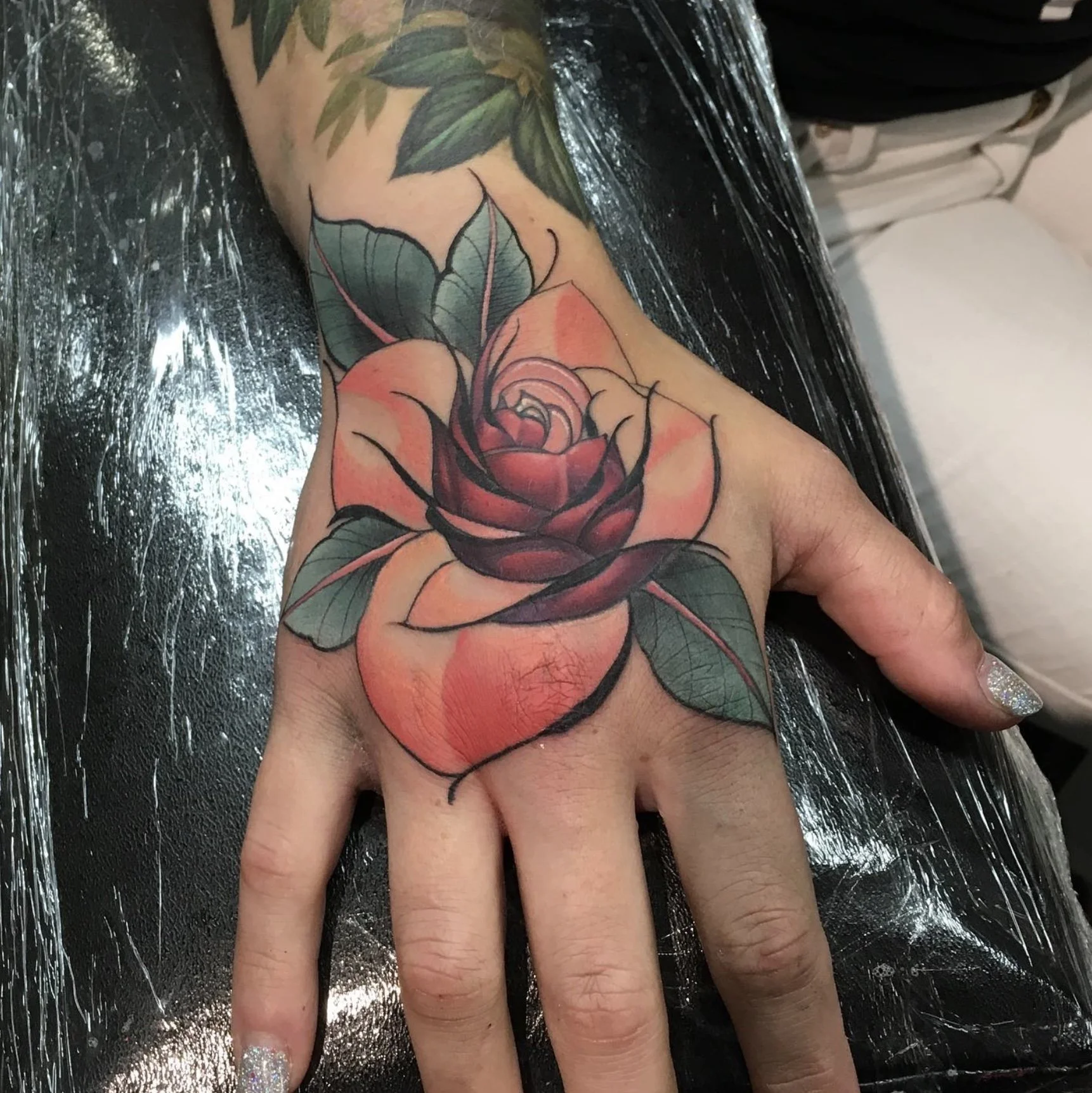 Newly completed colorful rose tattoo on hand with green leaves, lying on plastic sheet in a tattoo studio.