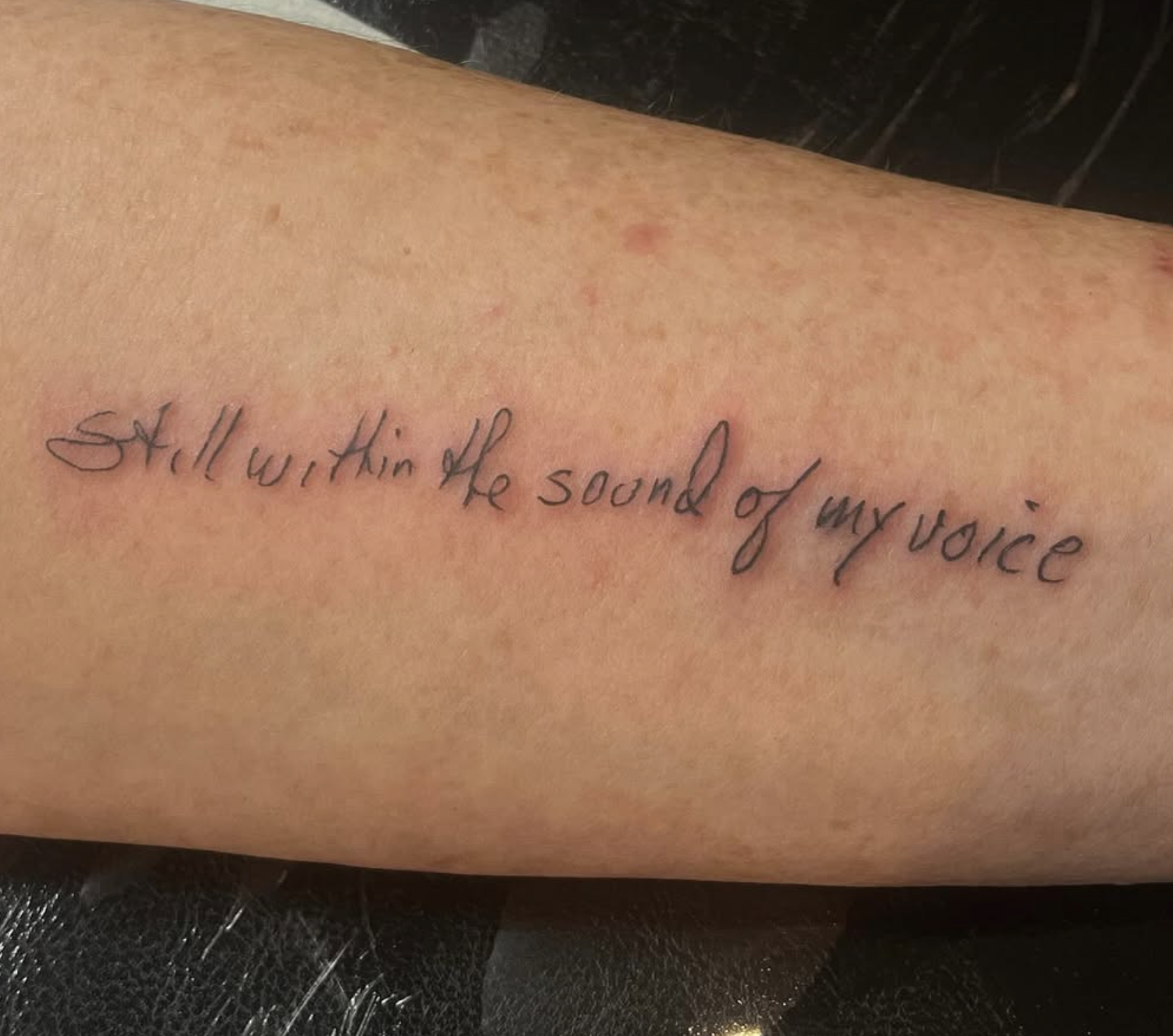 A person's arm with a tattoo of the phrase, 'Still worth it in the sound of my voice,' written in cursive black ink.