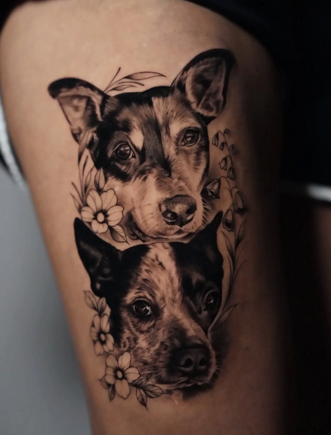 Black and gray tattoo of two dog heads, one above the other, surrounded by floral designs on skin.