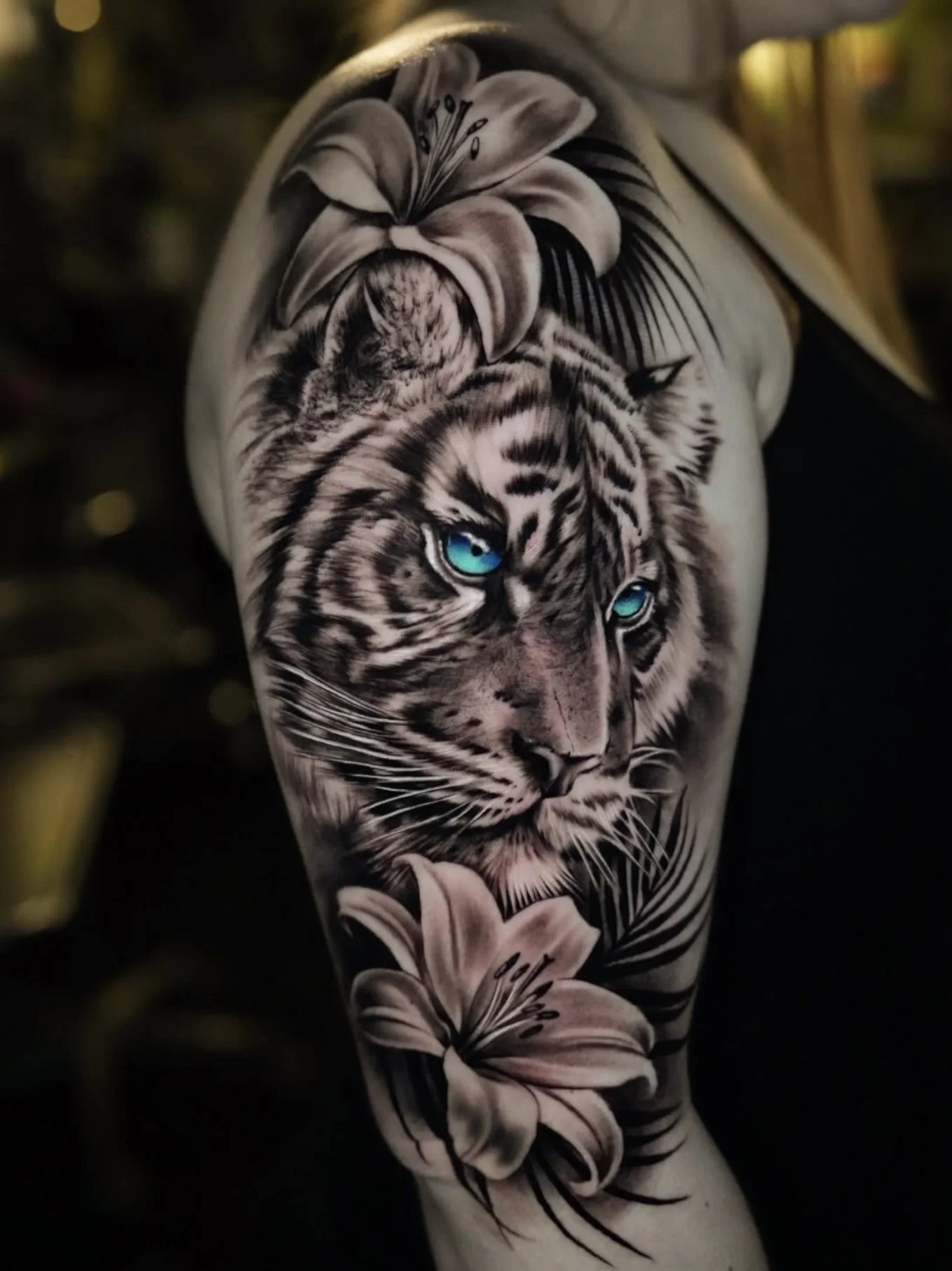 Black and gray realistic tiger face tattoo on person's arm, with blue eyes and surrounded by beige lilies.