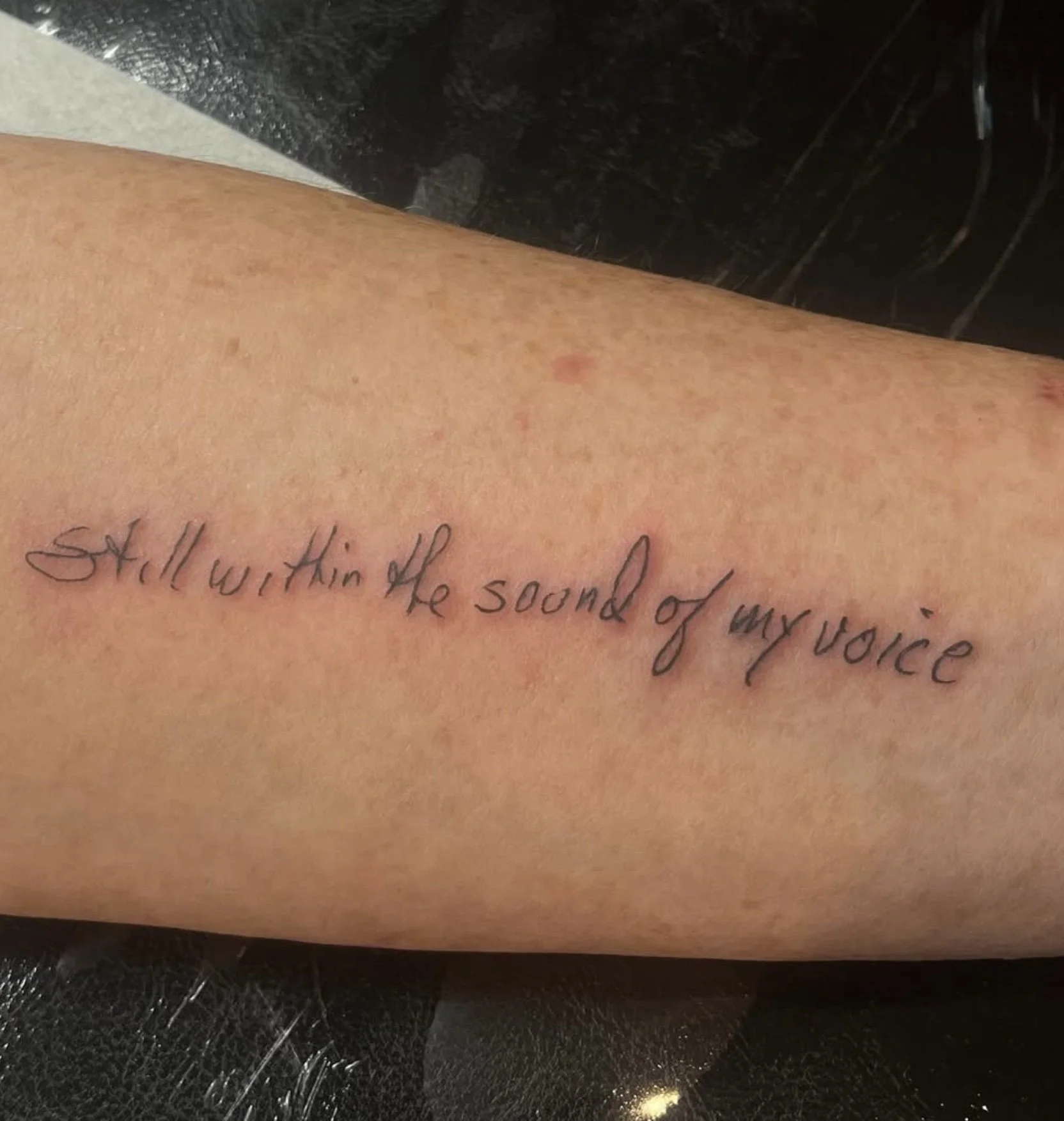 Close-up of a person's arm with a tattooed handwritten quote that reads 'Still working off the sound of my voice' on skin with some small red spots and blemishes, and a dark background.