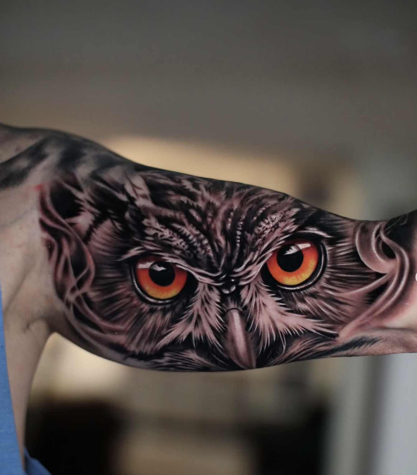 Tattoo of an owl's face with detailed feathers and piercing orange eyes on a person's arm.