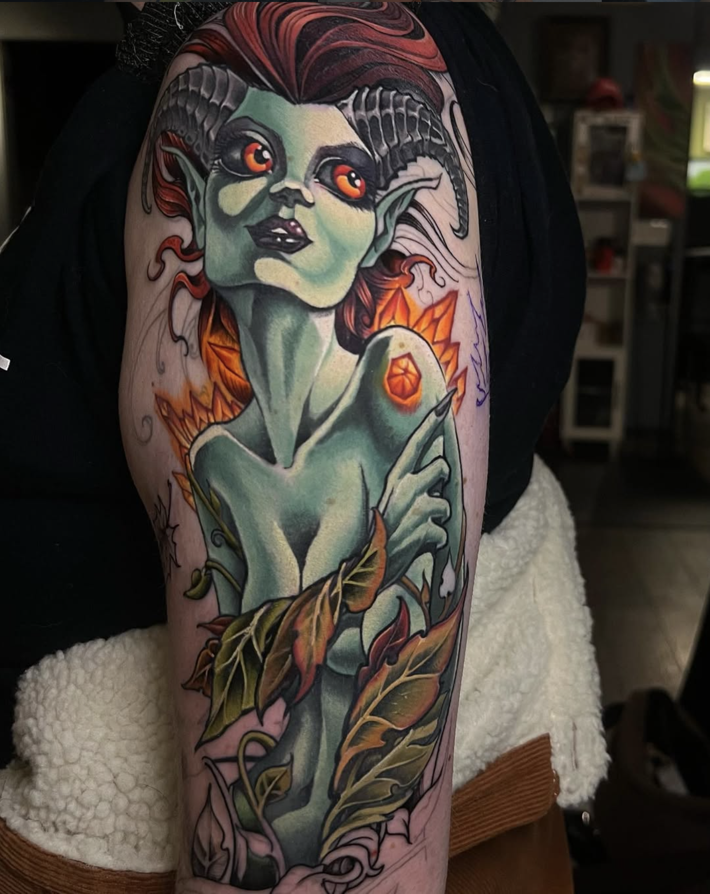Colorful tattoo of a mythical creature resembling a green-skinned woman with red eyes, horns, pointed ears, and surrounded by flames and leaves, on someone's upper arm.