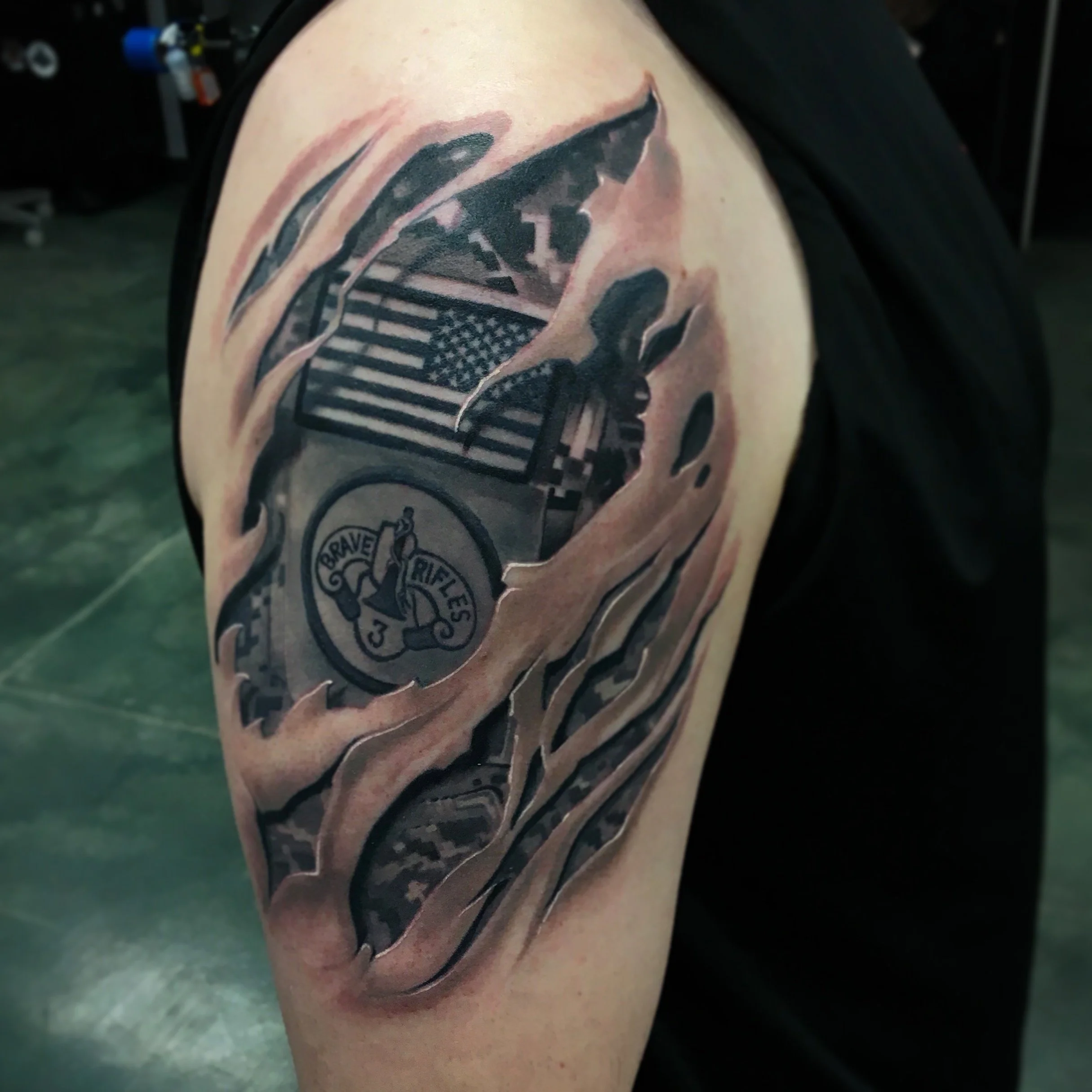 Black and gray tattoo on upper arm featuring an American flag, a shark, a badge reading 'BRAVE RIFLES 3', and abstract waves.