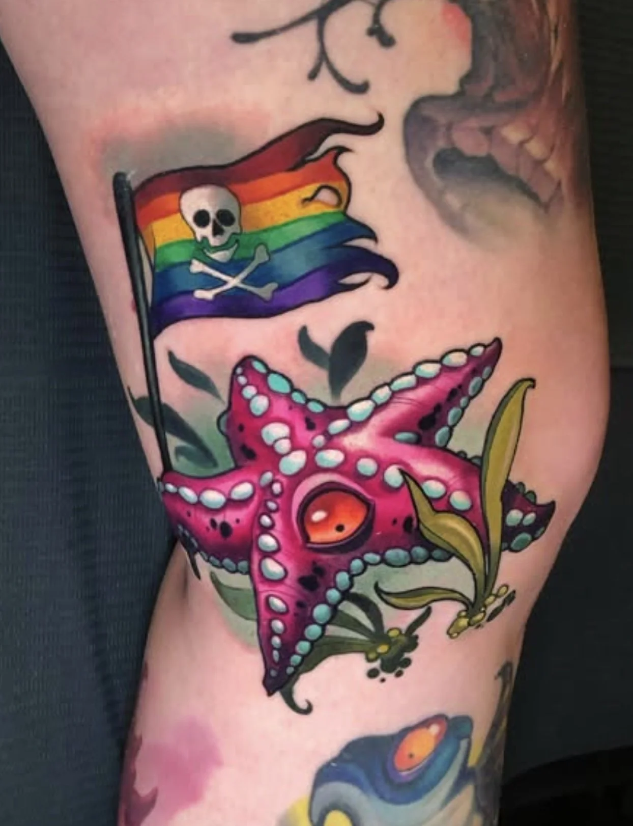Tattoo of a pink octopus with blue and white spots and an orange eye, along with a flag featuring a skull and crossbones in rainbow colors.